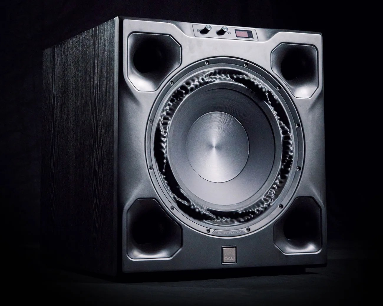 Black DALI subwoofer speaker with large central driver and four corner ports on a dark background.