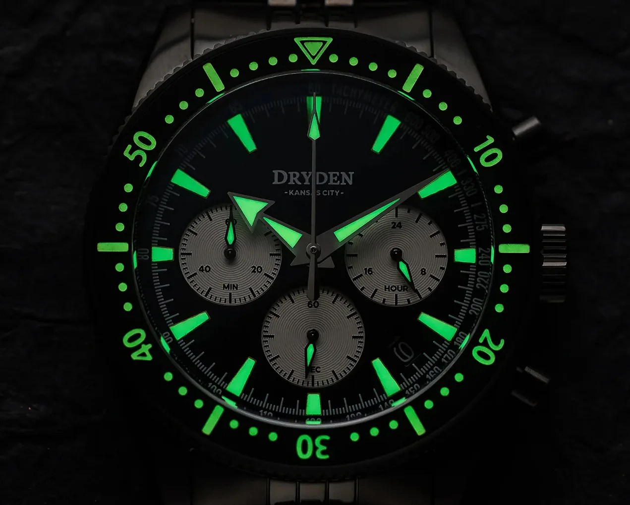 Black Dryden chronograph watch face with glowing green hour markers and hands in a dark setting.