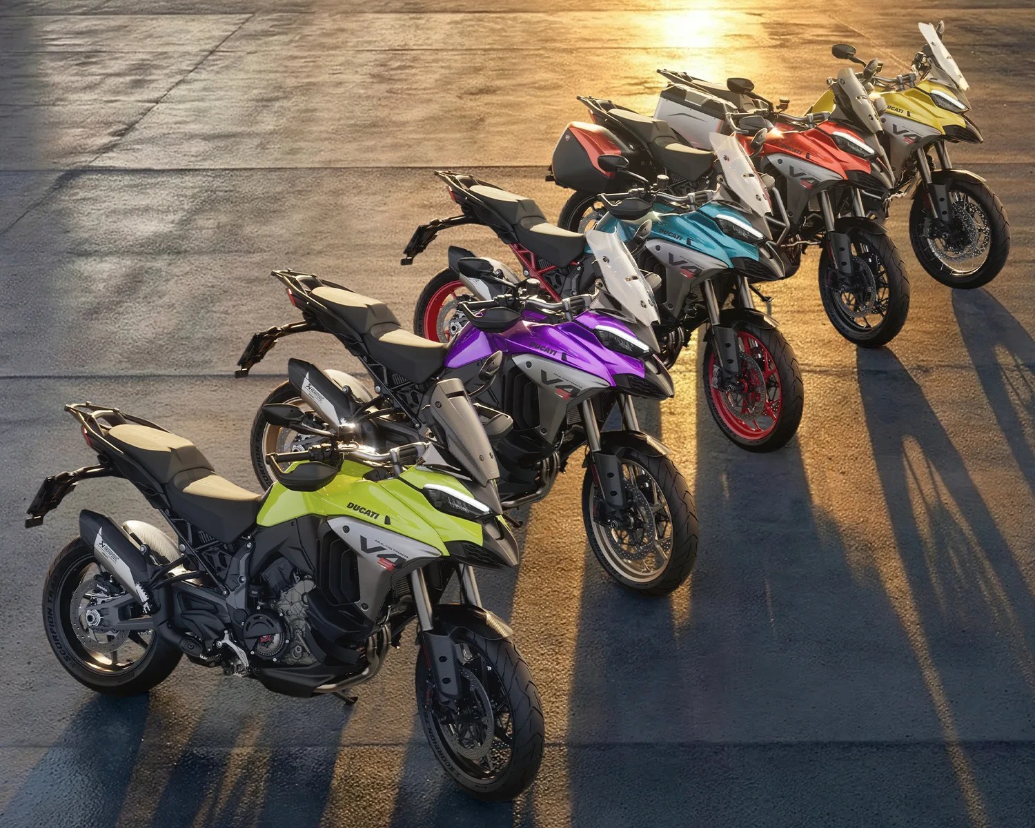 Five Ducati V4 motorcycles in yellow, purple, blue, orange, and yellow lined up on a sunlit pavement.