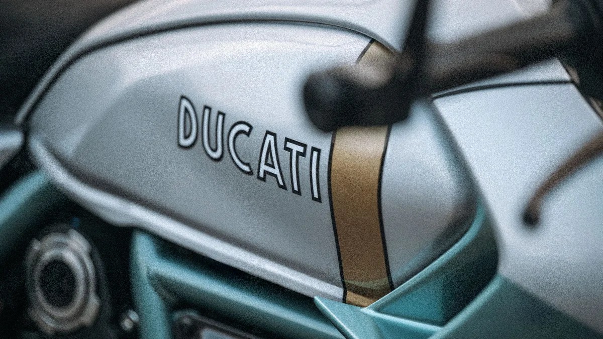 Close-up of a silver Ducati motorcycle fuel tank with a gold stripe and teal frame parts.