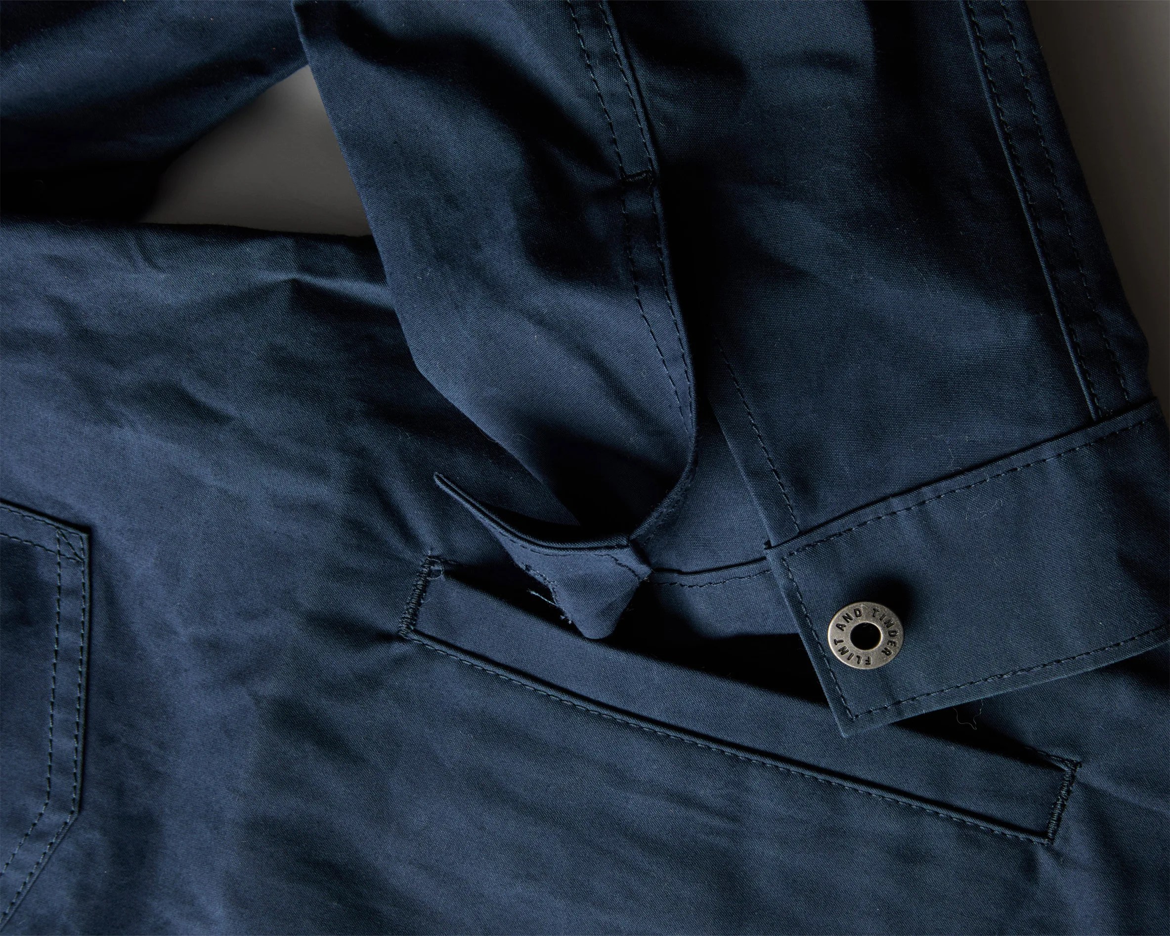 Close-up of navy blue fabric with a flap and a metal button engraved with "FLINT AND TINDER.