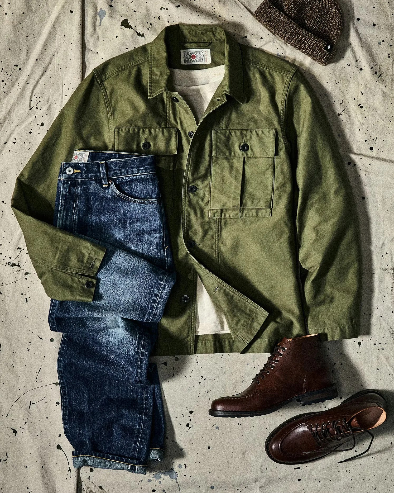 Olive green button-up jacket over a white shirt, paired with folded blue jeans, brown leather boots, and a brown knit beanie on a speckled fabric background.