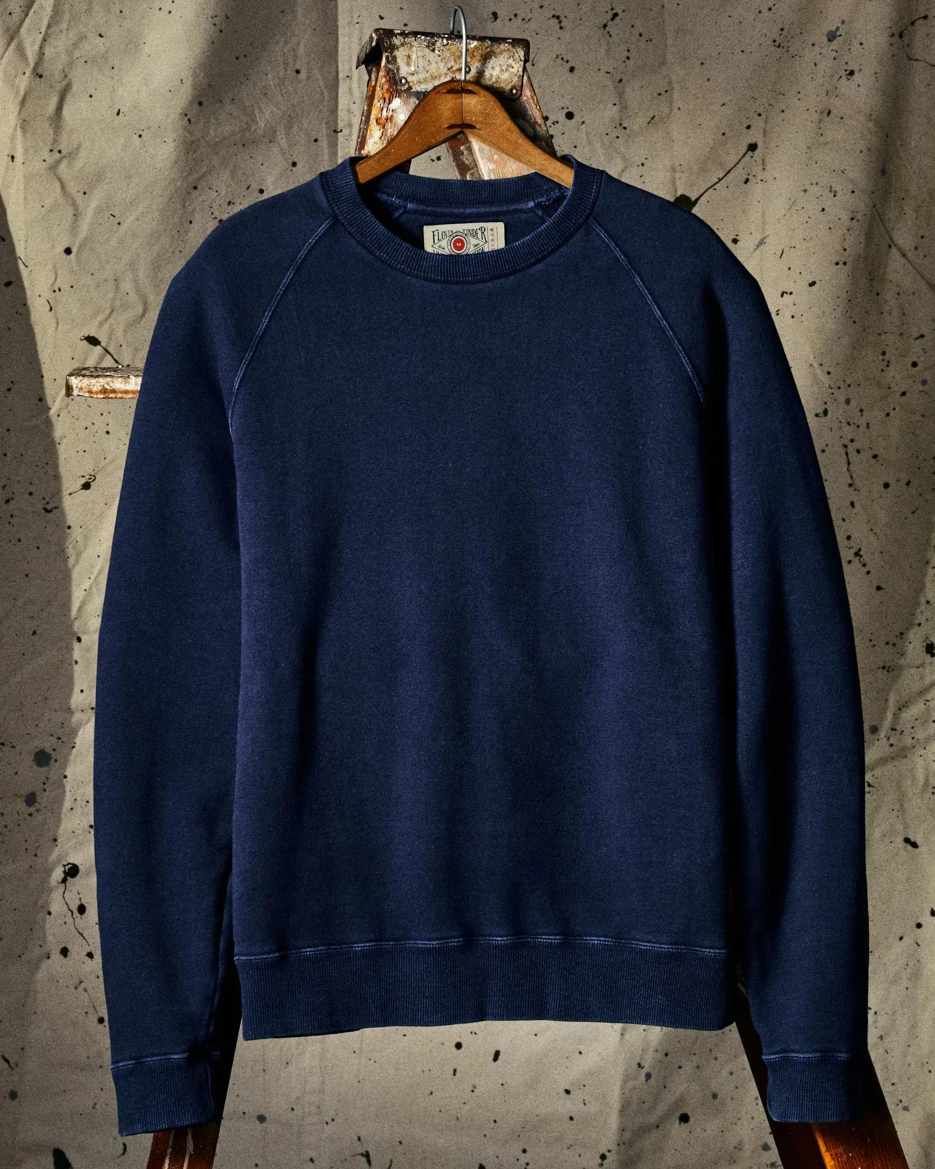 Navy blue crewneck sweatshirt hanging on a wooden hanger against a paint-splattered canvas backdrop.