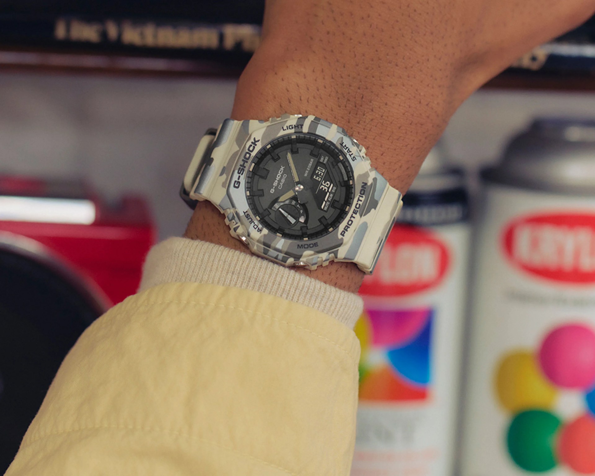 G-Shock Casio watch with gray camouflage band and black dial worn on a wrist with a beige jacket sleeve.