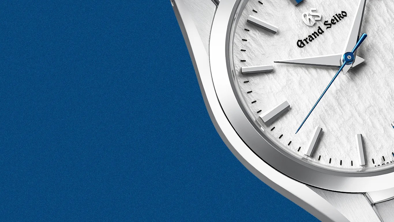Close-up of a Grand Seiko watch with a textured white dial, silver hour markers, and a blue second hand on a blue background.