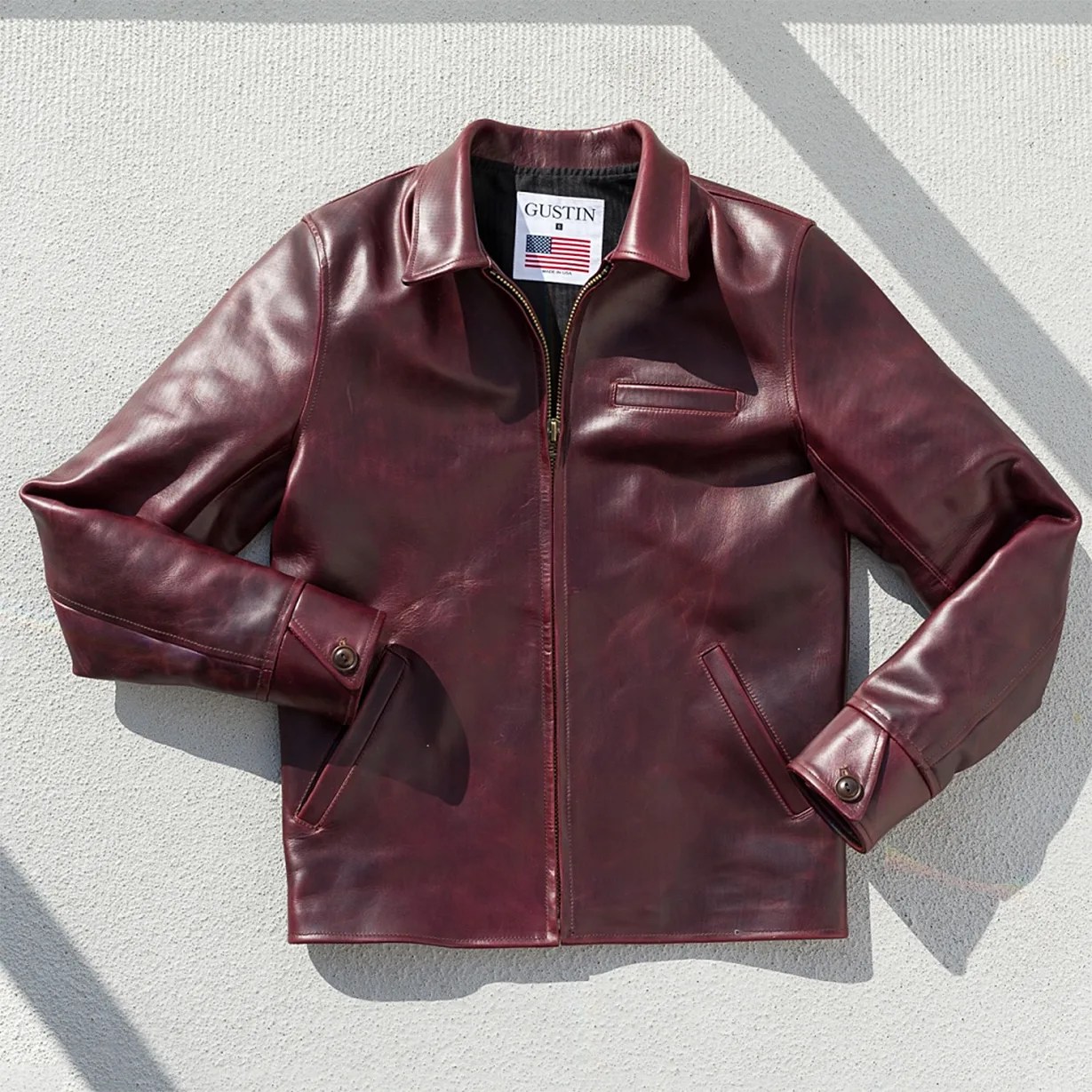 Burgundy leather jacket with a front zipper, buttoned cuffs, and a chest pocket, laid flat on a textured surface.