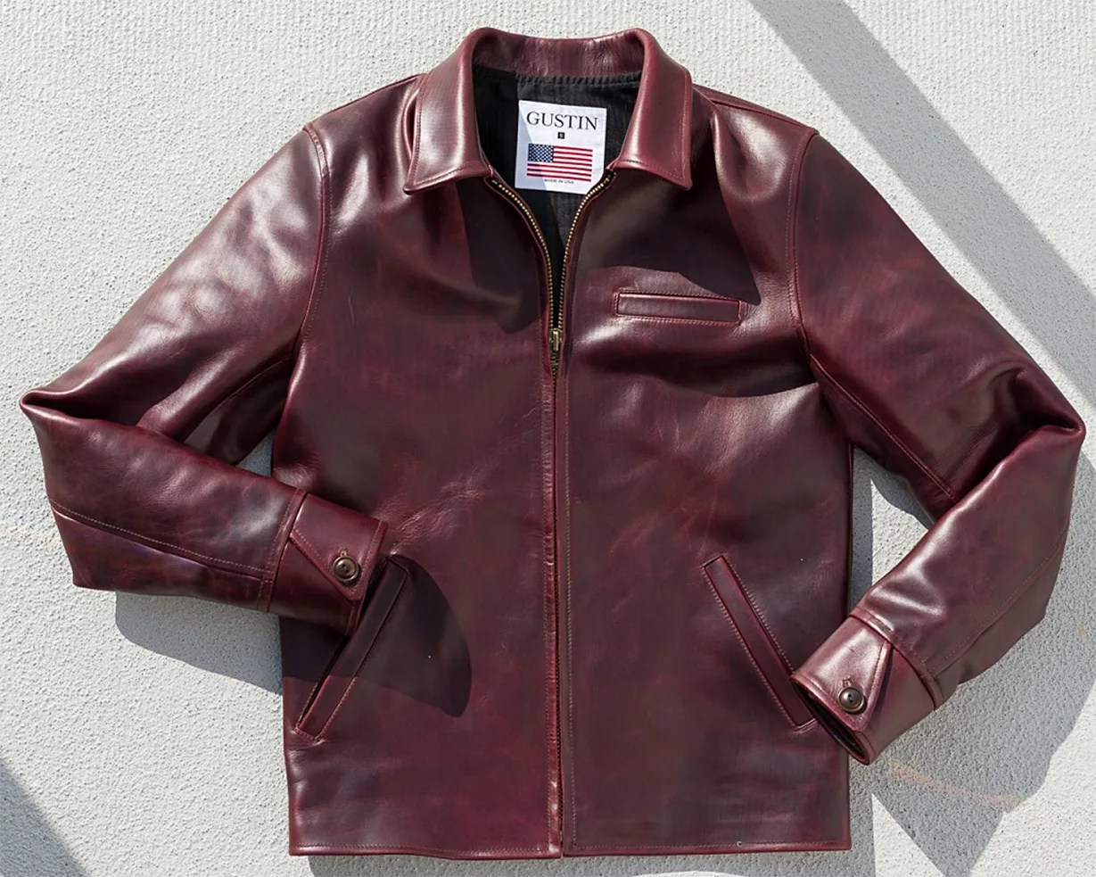 Burgundy leather jacket with front zipper, buttoned cuffs, and chest pocket laid flat on a light textured surface.