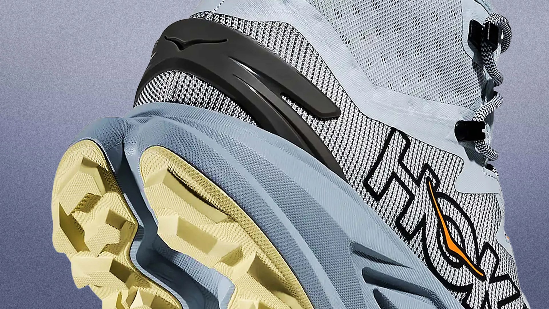 Close-up of a light blue and beige trail running shoe with black and orange "HOKA" branding and rugged sole.