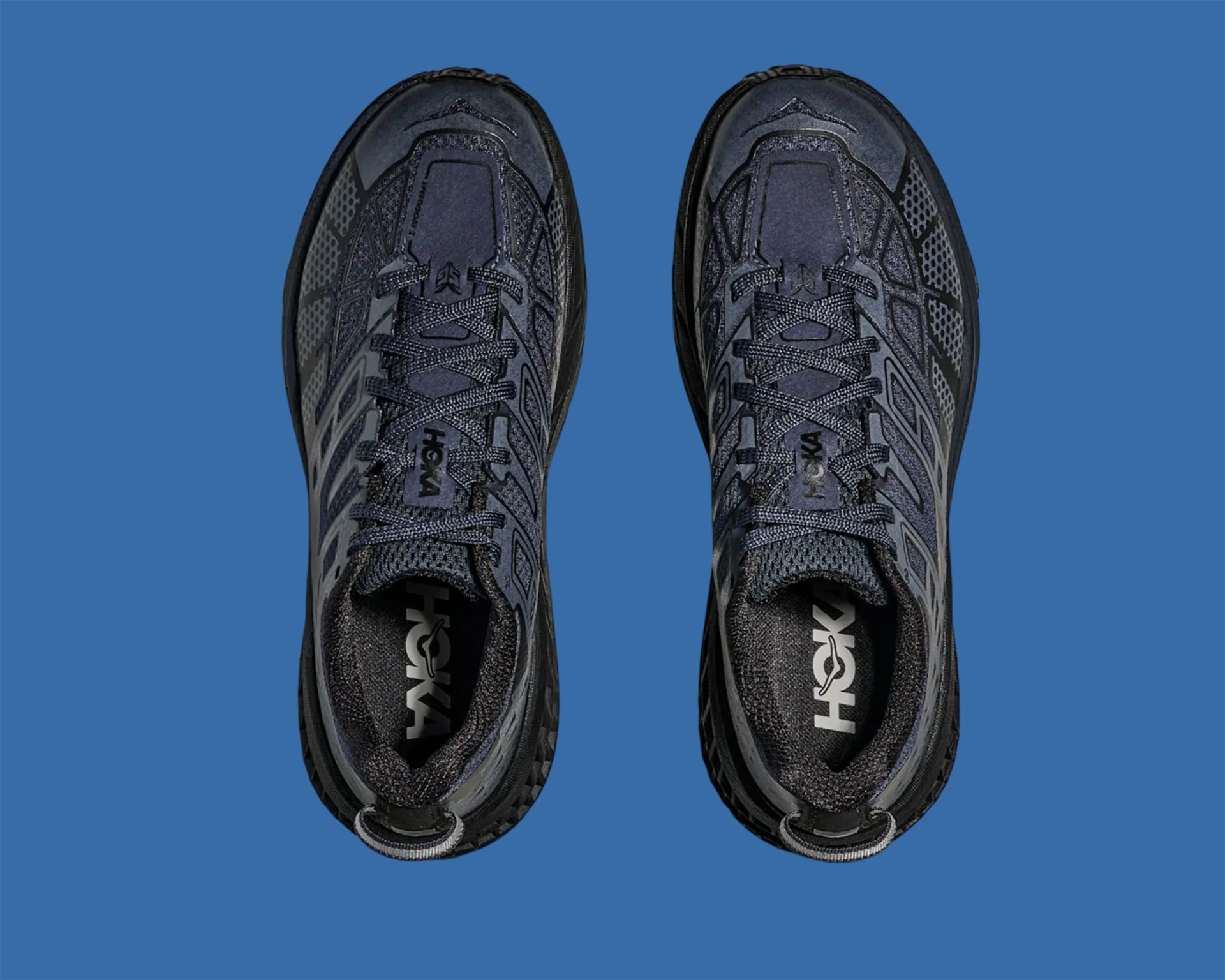 Pair of black and dark gray Hoka running shoes viewed from above on a blue background.