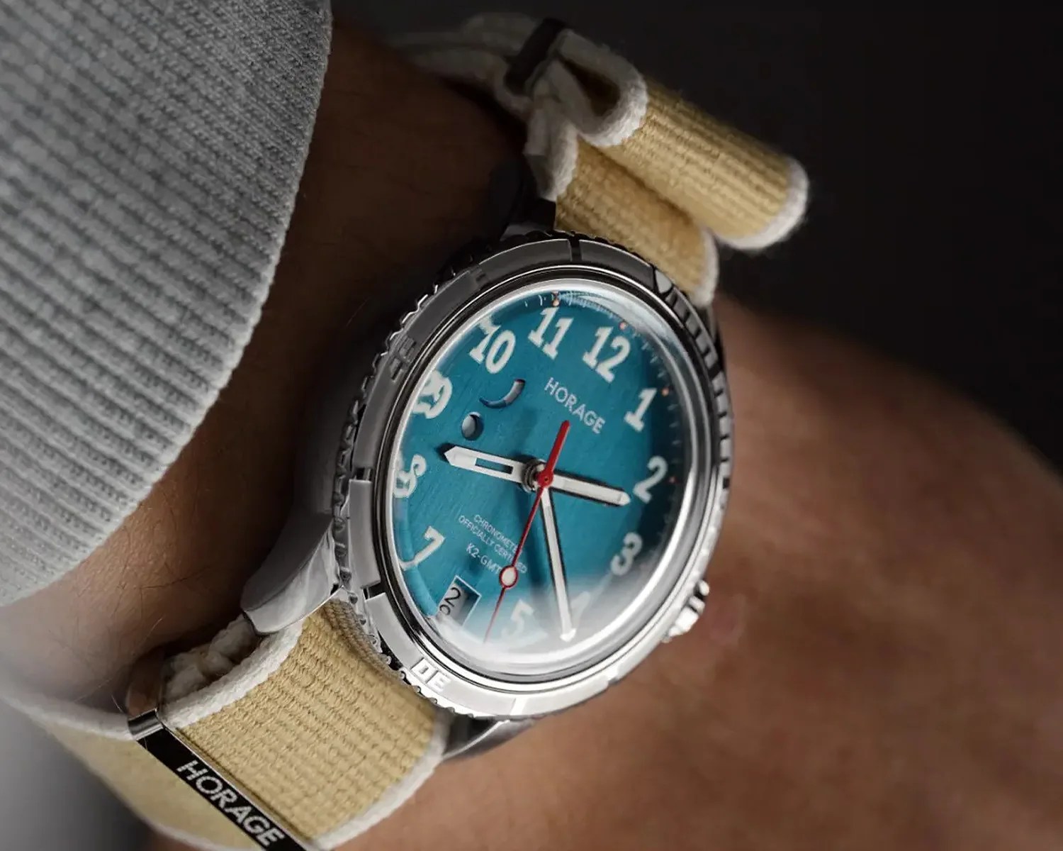 Silver wristwatch with a blue dial, white numerals, red second hand, and beige fabric strap worn on a wrist.