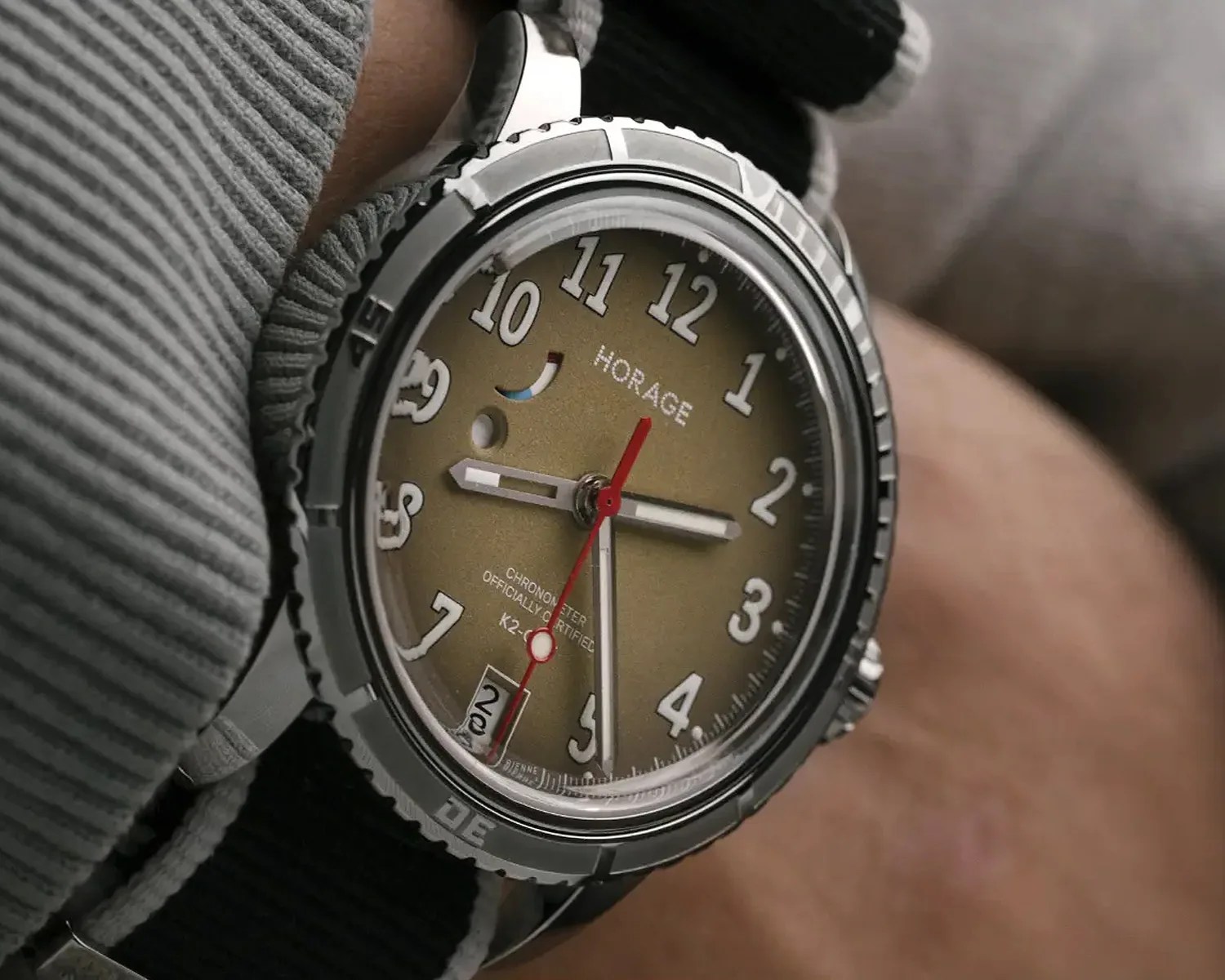 Close-up of a wristwatch with a metallic bezel, olive-green dial, white numerals, red second hand, and black fabric strap.