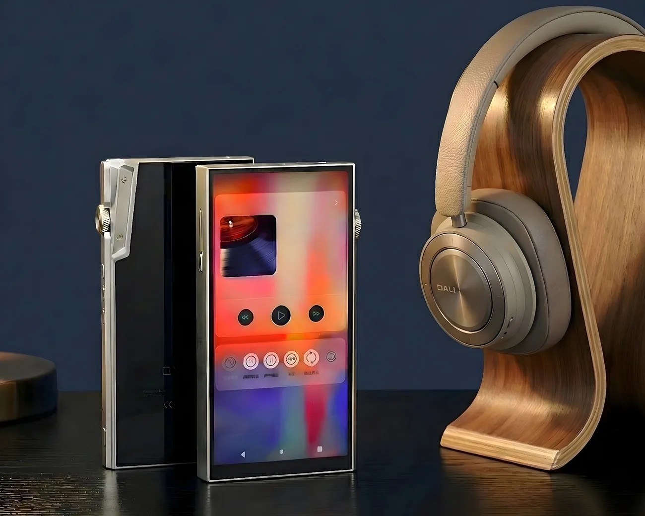 Two rectangular digital music players with metal frames and screens, next to beige over-ear headphones on a curved wooden stand.