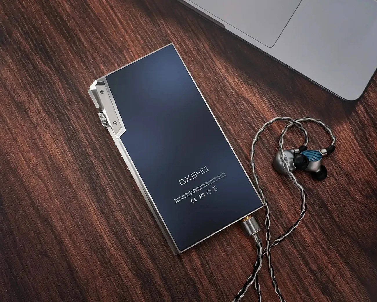 Rectangular digital audio player with a dark blue front and silver edges connected to silver and black in-ear monitors on a wooden surface.