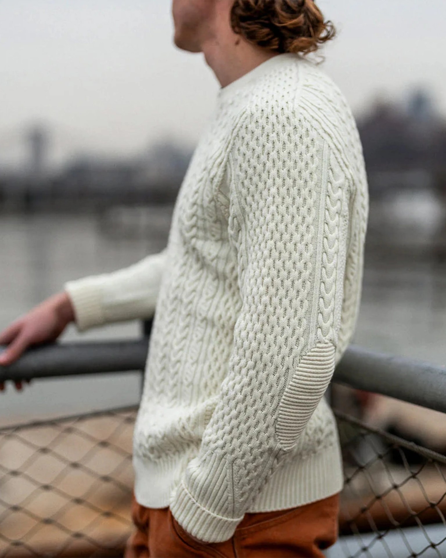 Cream-colored cable knit sweater with elbow patches worn by a person leaning on a railing.
