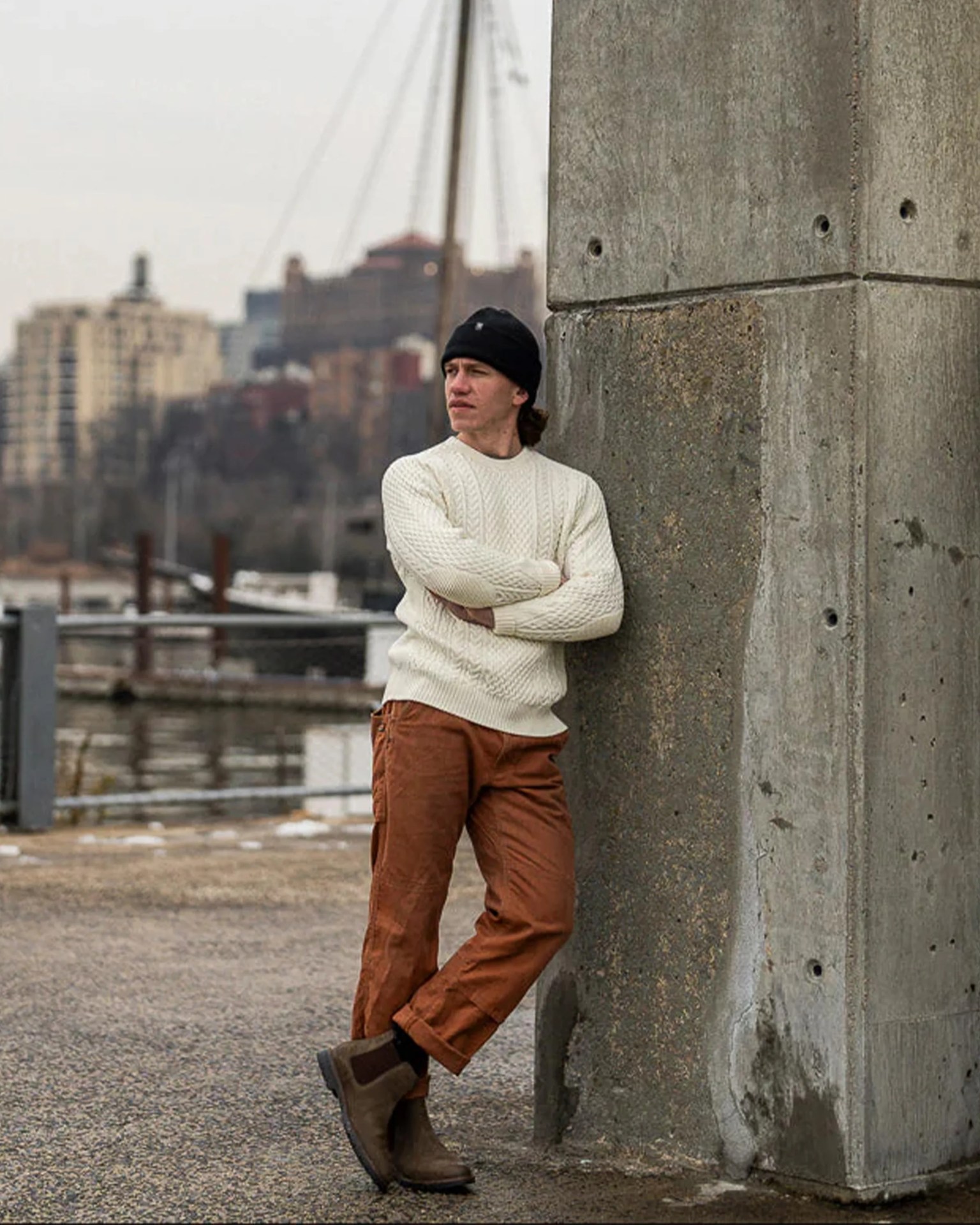 Person wearing a cream cable-knit sweater, rust-colored pants, brown boots, and a black beanie leaning against a concrete pillar.