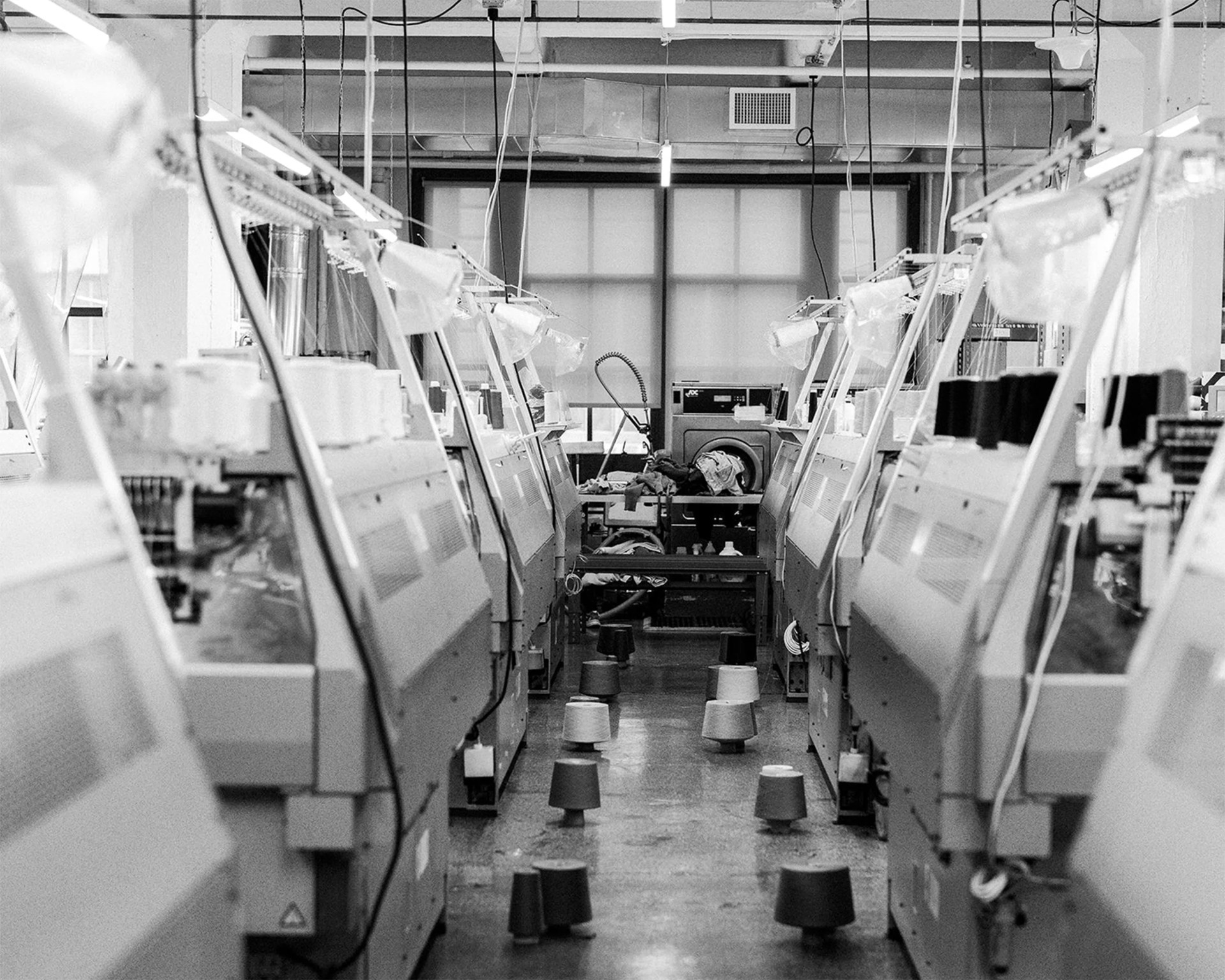 Black and white photo of a textile factory with rows of knitting machines and yarn cones scattered on the floor.