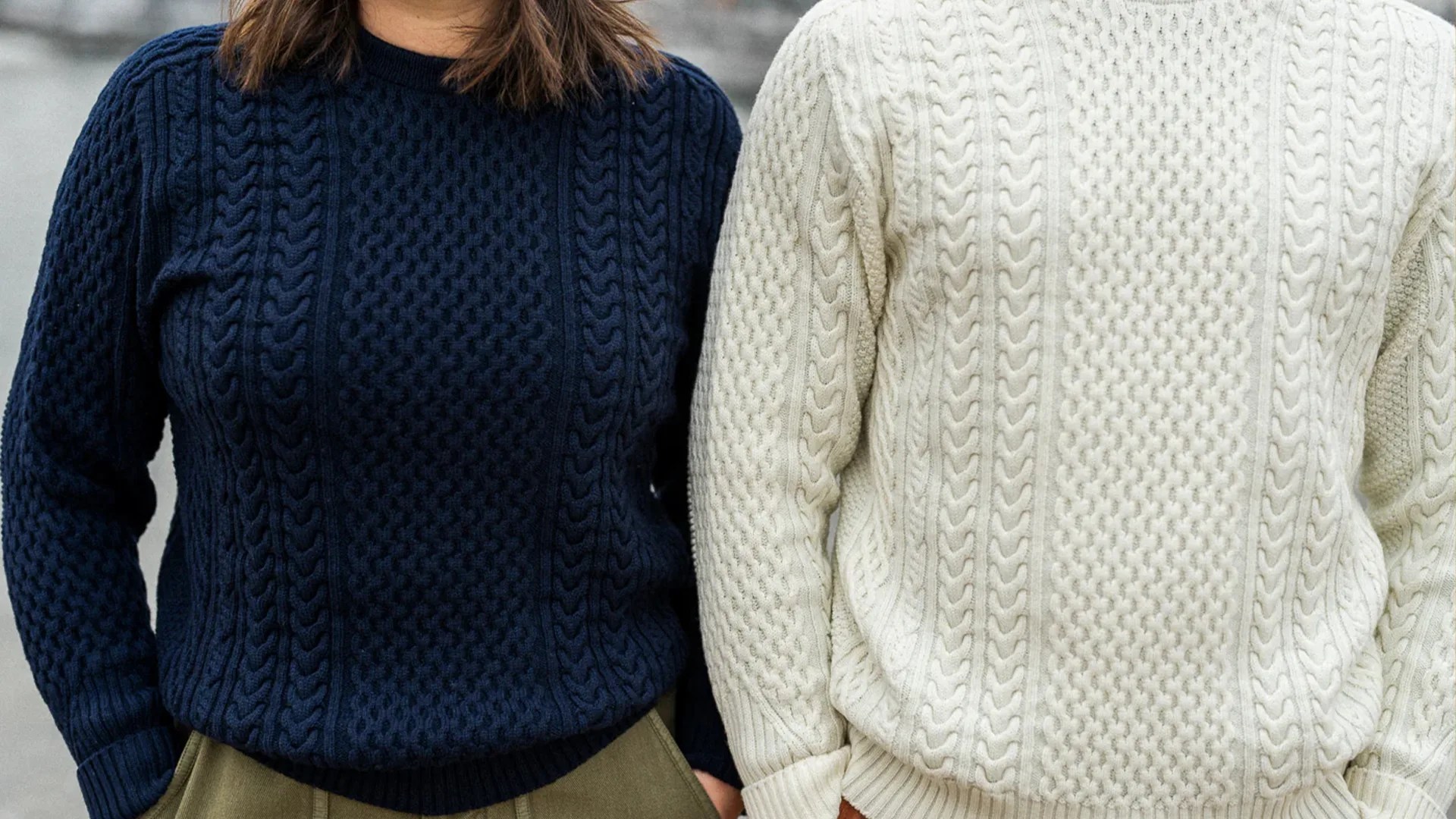 Two people wearing cable-knit sweaters, one navy blue and one cream, both with textured patterns.