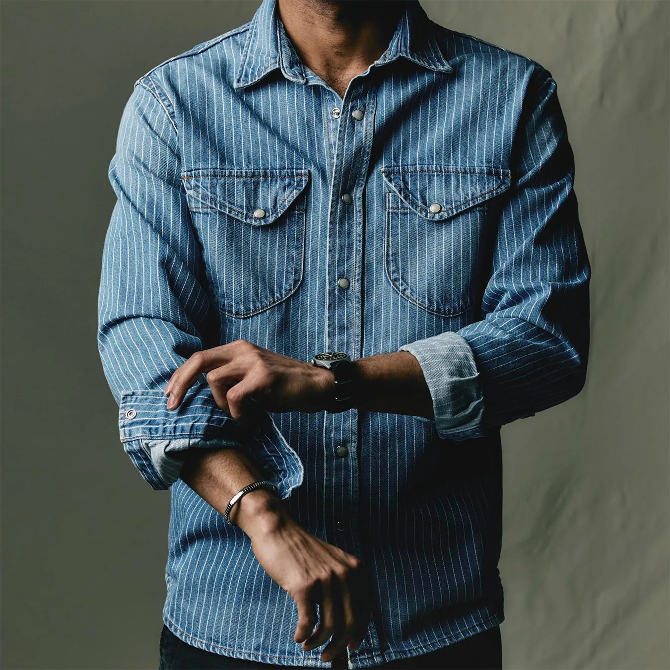 Blue denim shirt with white vertical stripes and buttoned chest pockets worn by a person rolling up the sleeves.