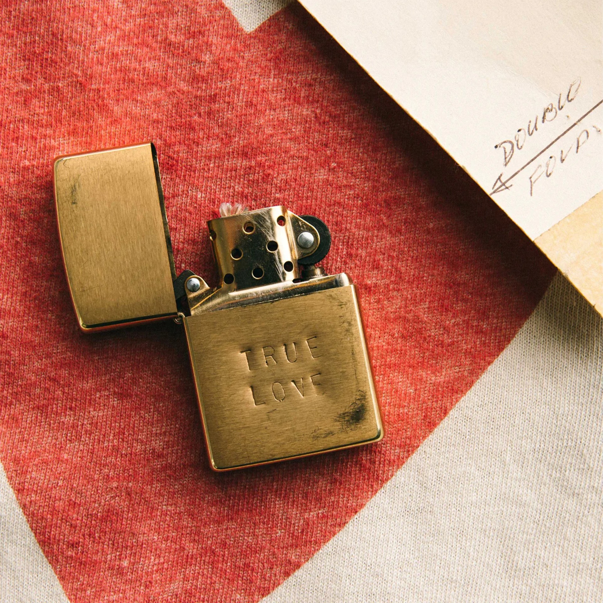 Brass lighter with "TRUE LOVE" engraved on the front, placed on red and white fabric next to a paper corner.