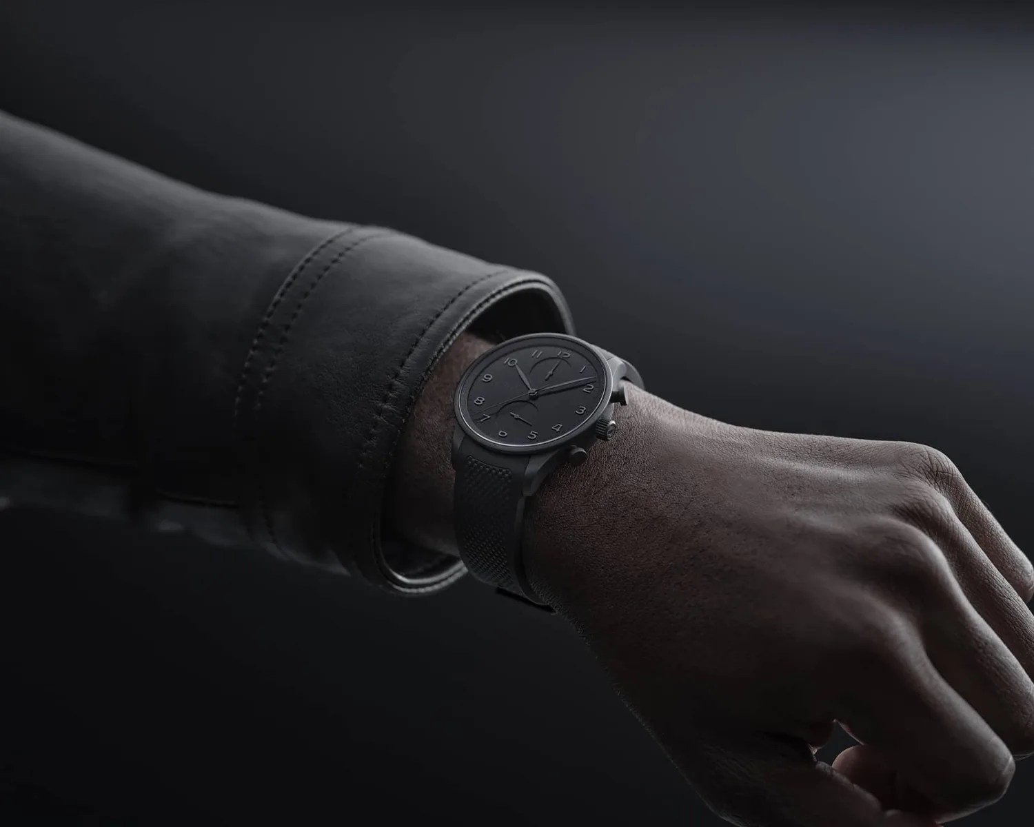 Black wristwatch with textured strap worn on a wrist with a black leather jacket sleeve.