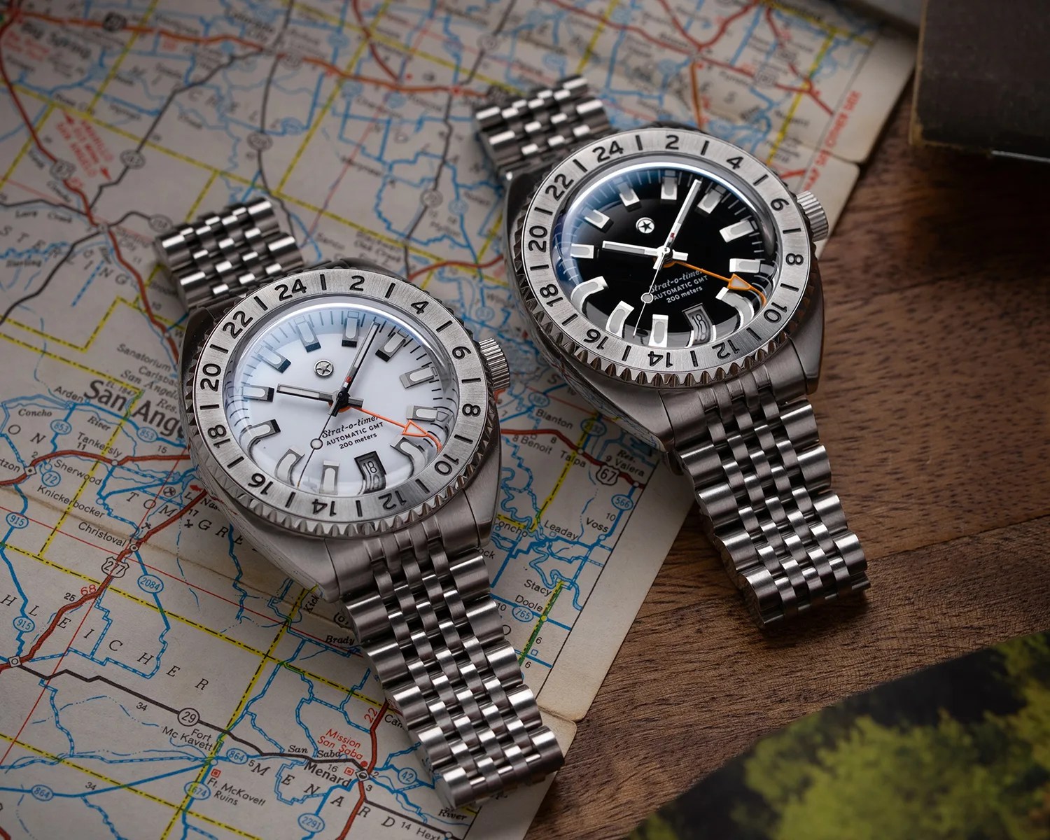Two stainless steel wristwatches with black and white dials and 24-hour bezels on a map and wooden surface.