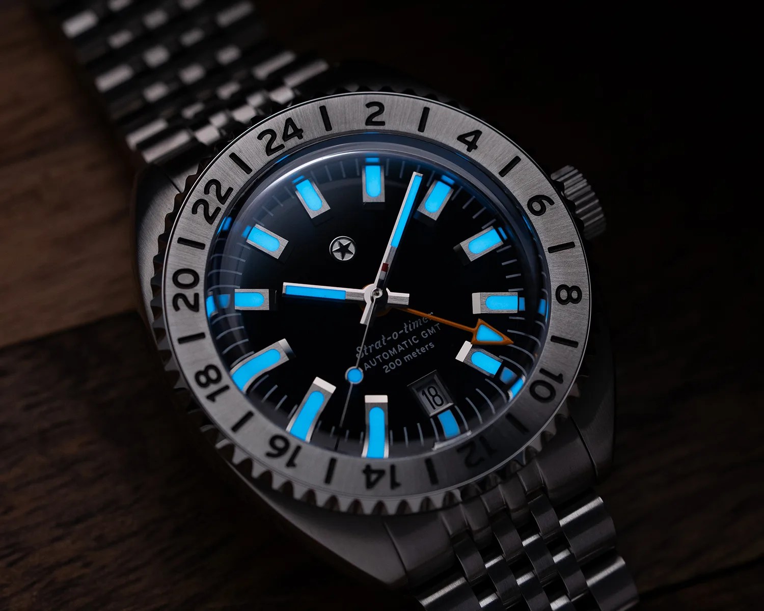 Close-up of a stainless steel wristwatch with luminous blue hour markers and hands, a 24-hour bezel, and a date window at 4 o'clock.