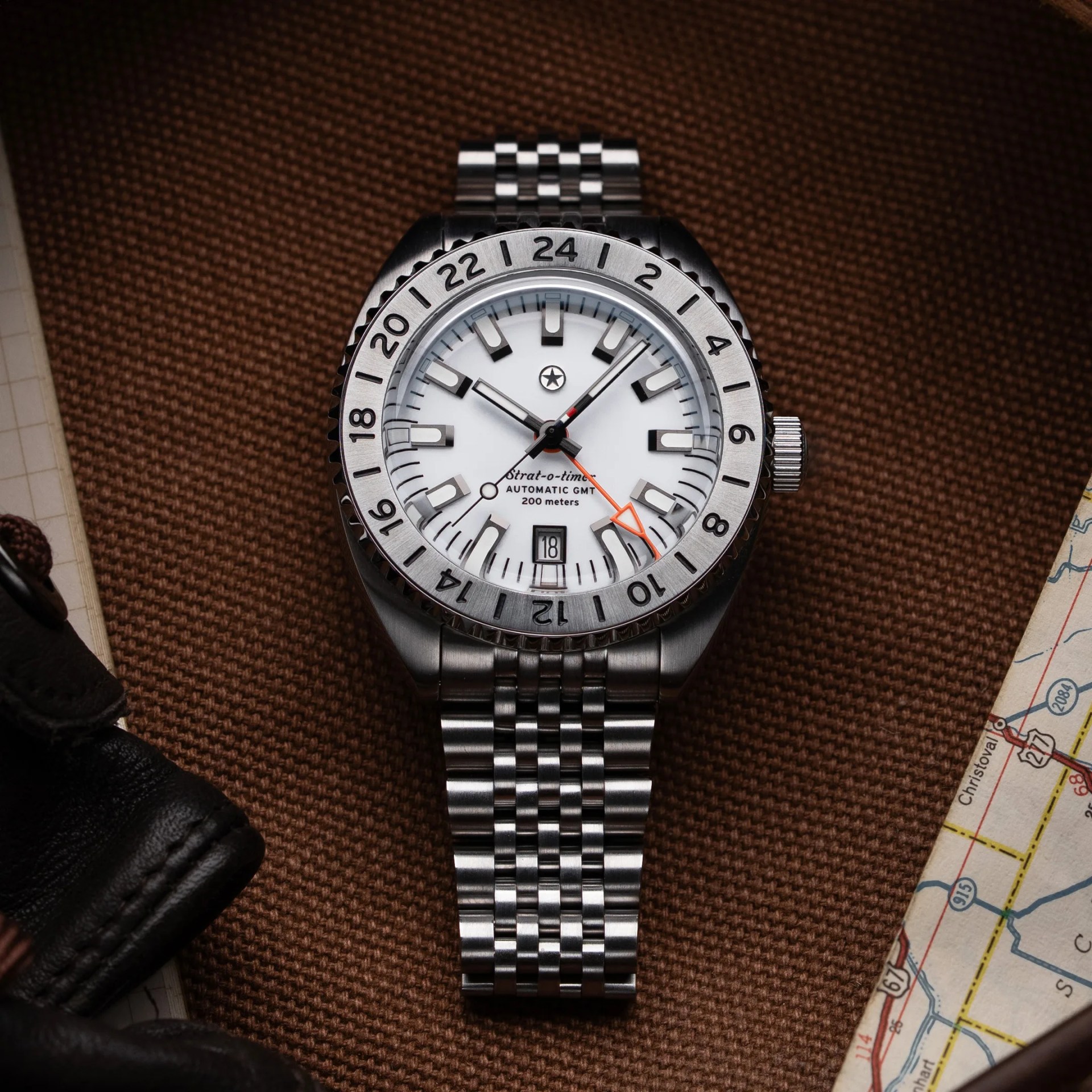 Stainless steel automatic GMT wristwatch with white dial, black hour markers, and orange GMT hand on brown textured surface.