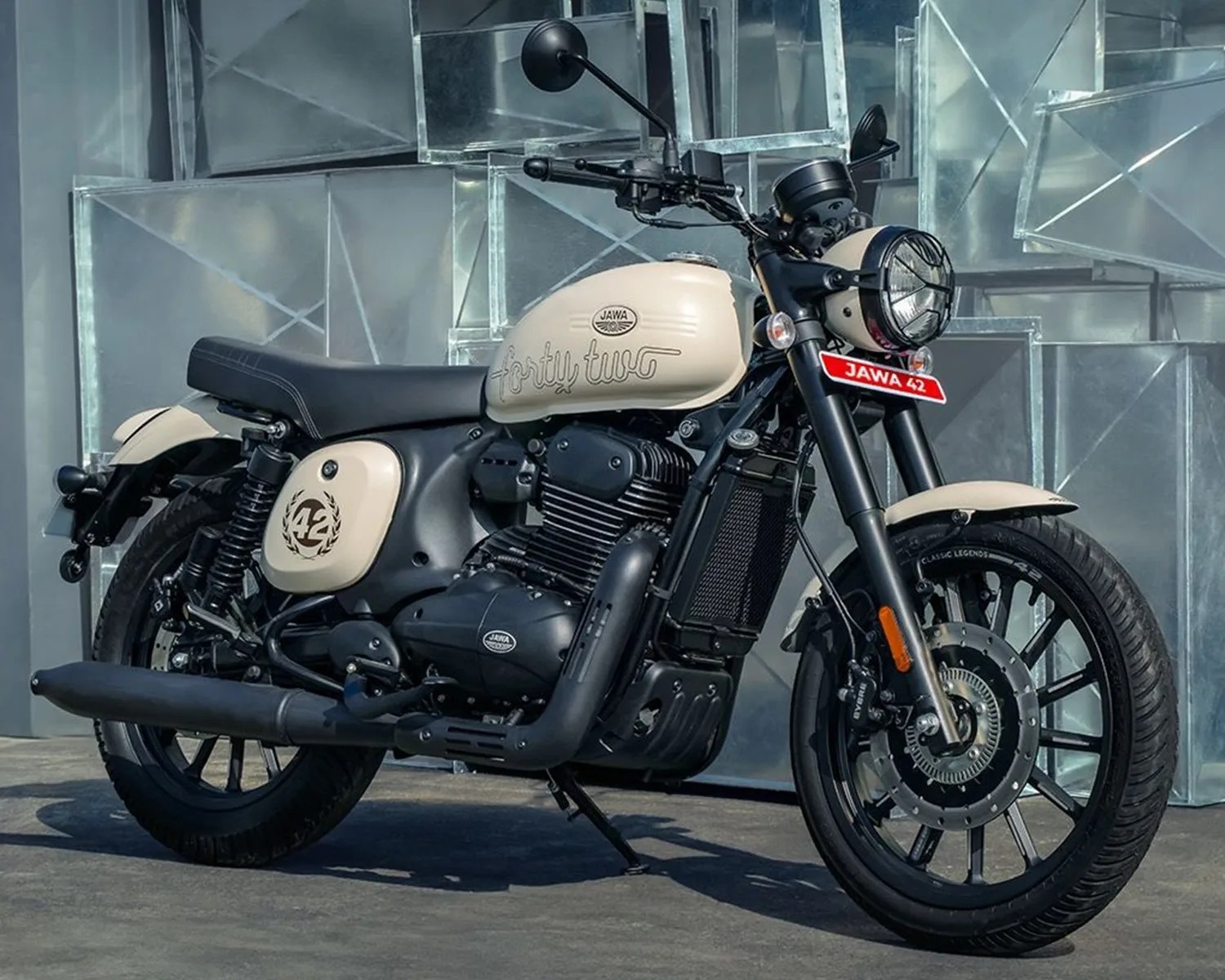 Cream and black Jawa 42 motorcycle parked on a concrete surface with a metallic geometric background.
