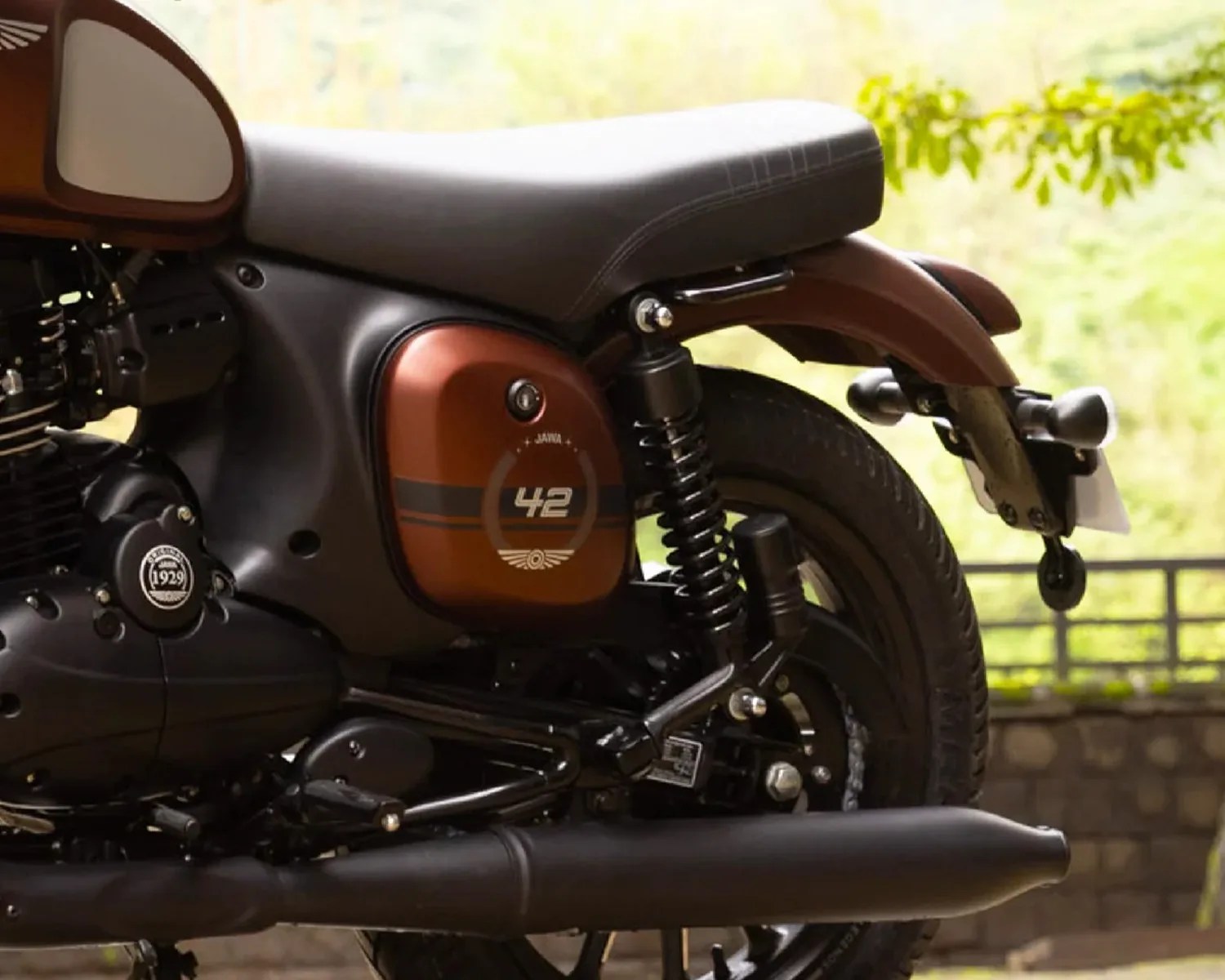 Close-up of the rear section of a brown and black Jawa 42 motorcycle showing the seat, rear suspension, exhaust, and rear tire.