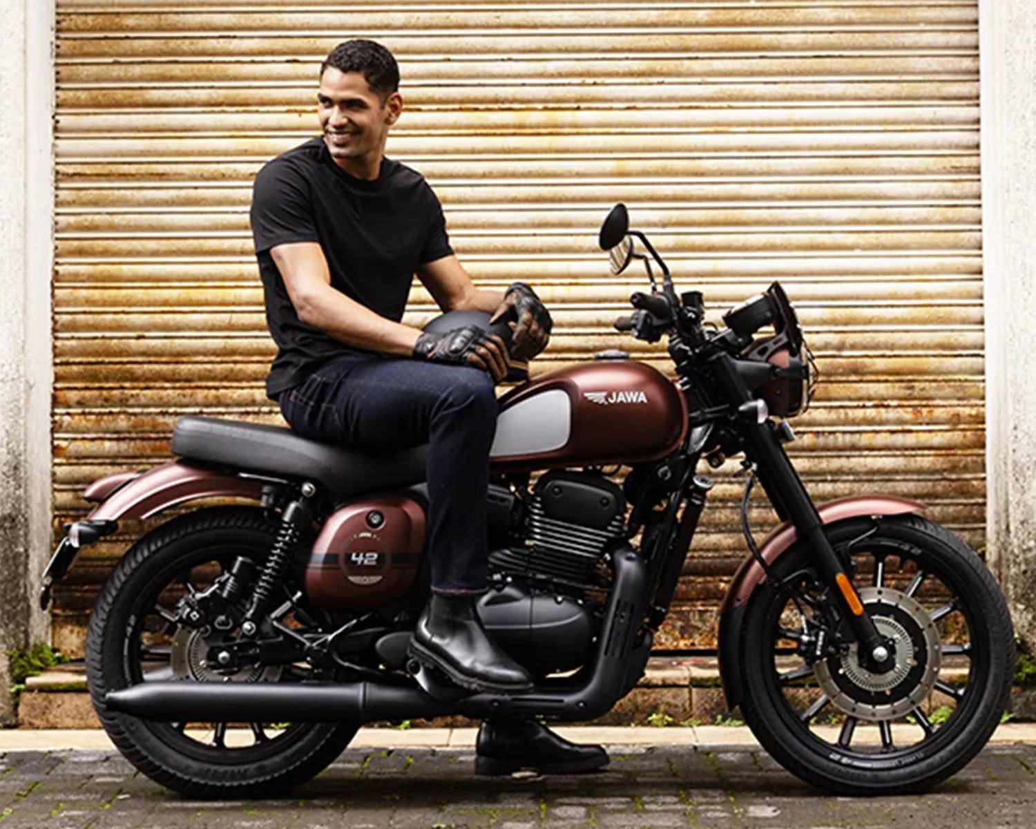 Man in black shirt and gloves sitting on a matte brown Jawa motorcycle against a rusty metal shutter.