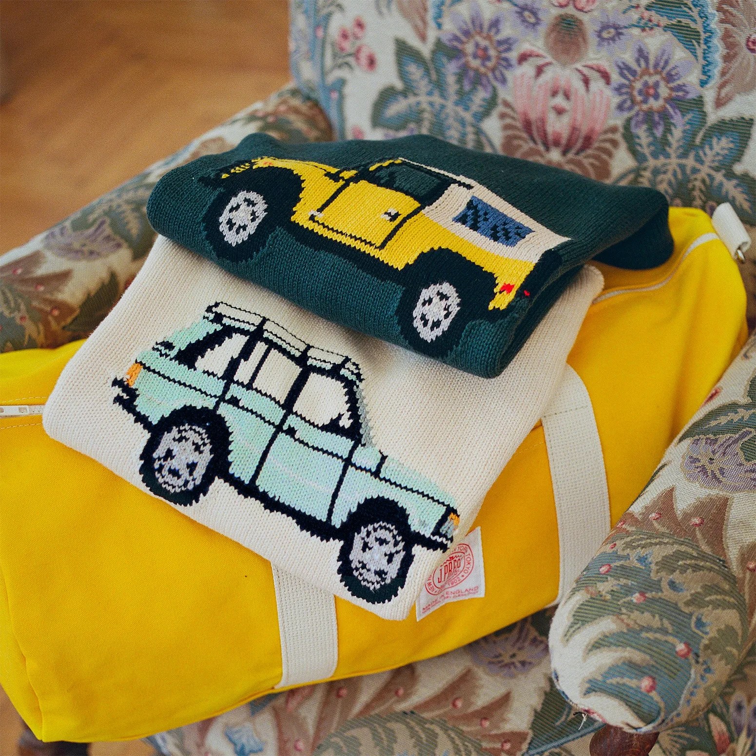Two folded knit sweaters with car designs, one yellow and black on dark green and one light blue and black on cream, on a yellow bag on a floral armchair.