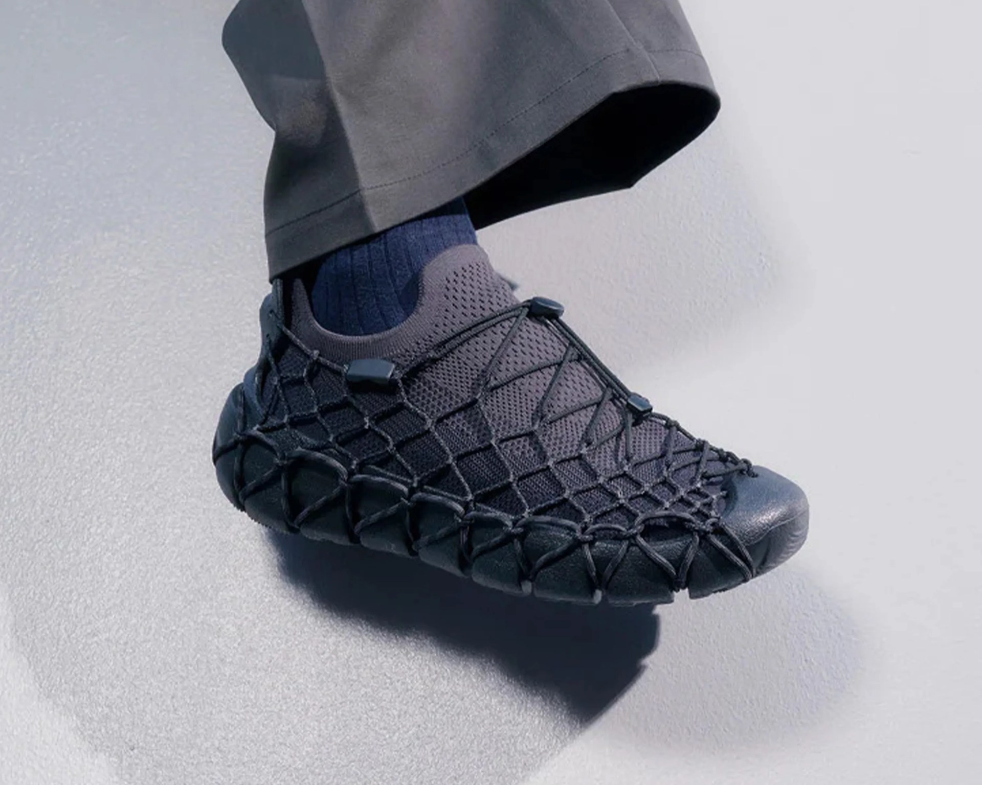 Black slip-on sneaker with mesh upper and an outer net-like cord design worn with dark pants and socks.