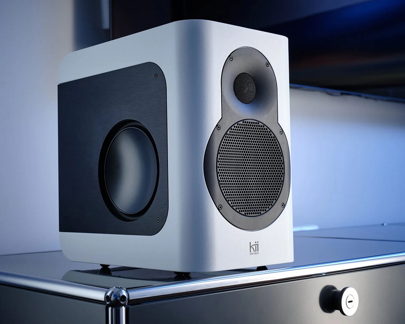 White and black Kii Seven speaker with front and side drivers on a metal surface.