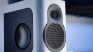 White speaker with black circular mesh grille and side bass radiator on a blurred background.