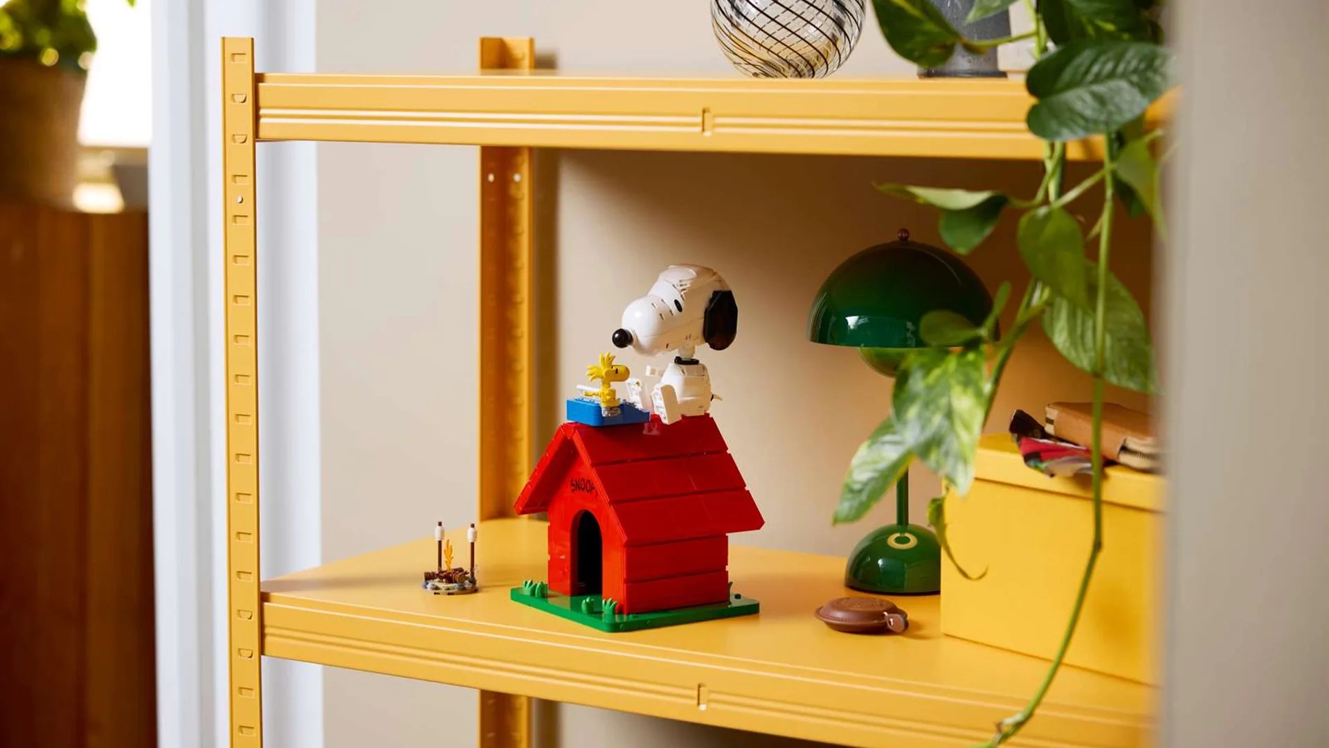 Red LEGO Snoopy doghouse with Snoopy and Woodstock figures on a yellow metal shelf next to a green lamp and a yellow box.