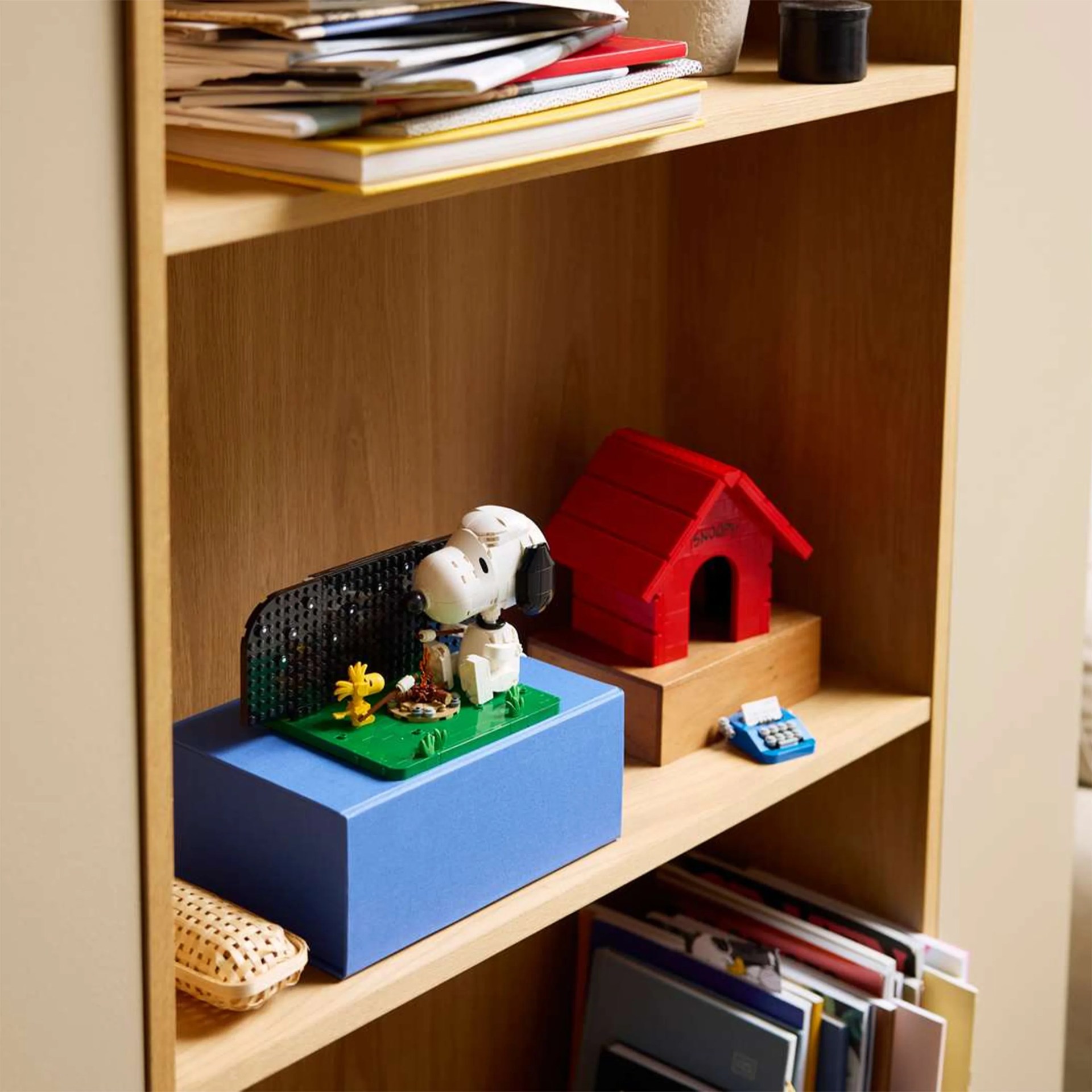 Wooden shelf with a Snoopy and Woodstock LEGO set on a blue box, a red Snoopy doghouse model, and stacked books.