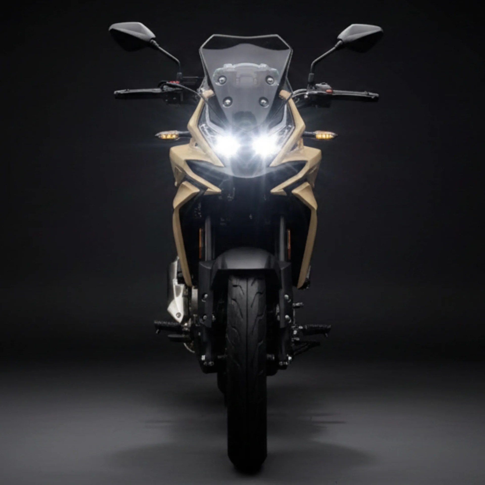 Front view of a beige and black motorcycle with headlights on against a dark background.