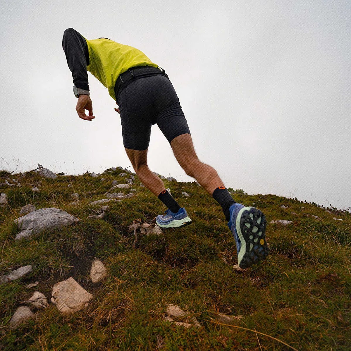 Person wearing blue trail running shoes and black shorts running uphill on rocky grass terrain.