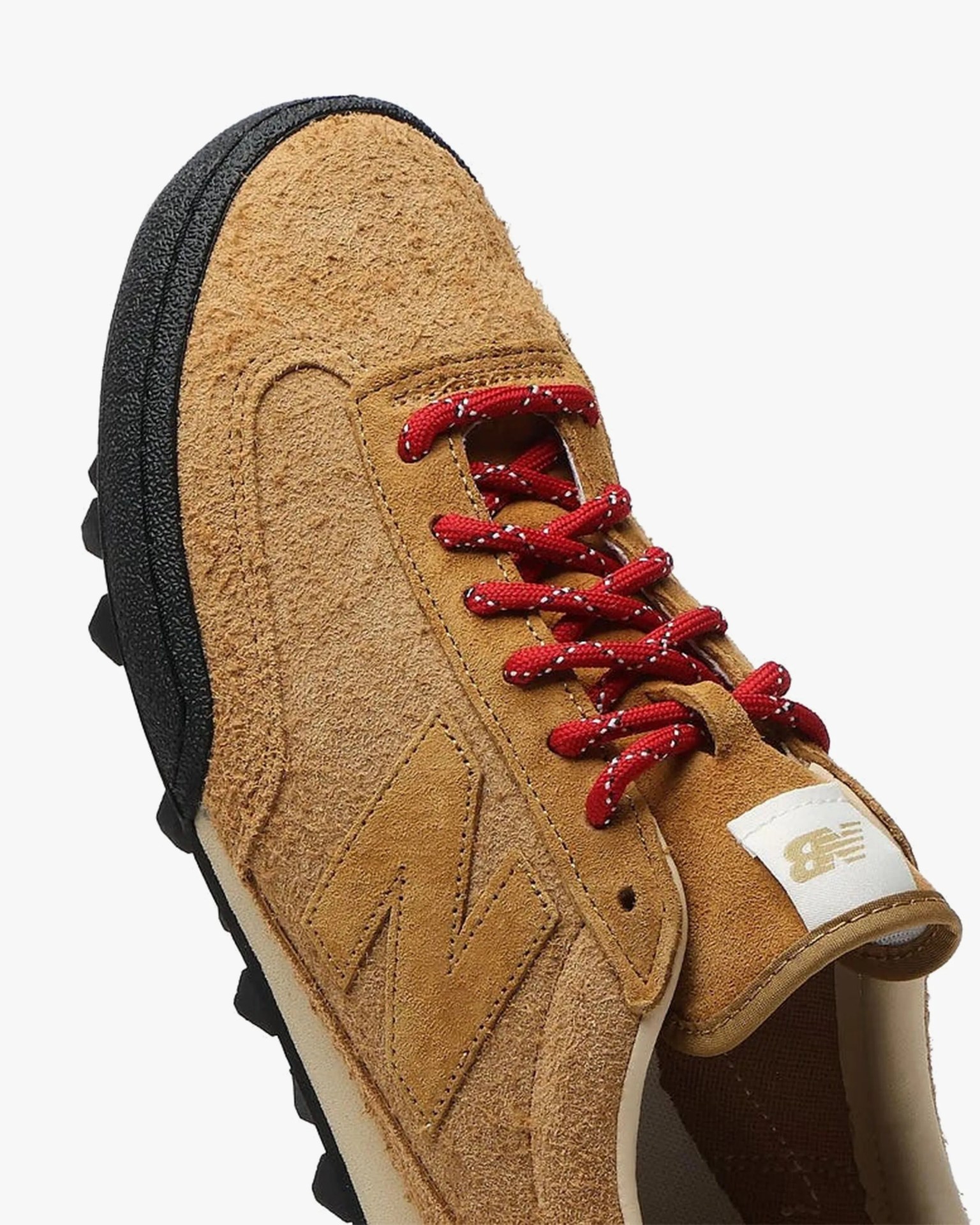 Tan suede New Balance sneaker with red and white speckled laces and black textured sole.