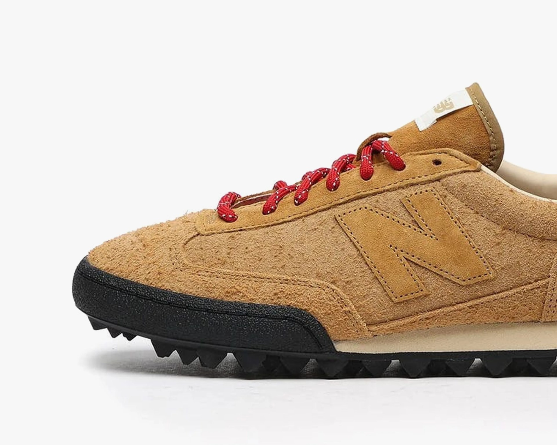Tan suede New Balance sneaker with red laces and black textured sole.