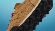 Brown suede sneaker with red laces and black studded sole against a blue gradient background.