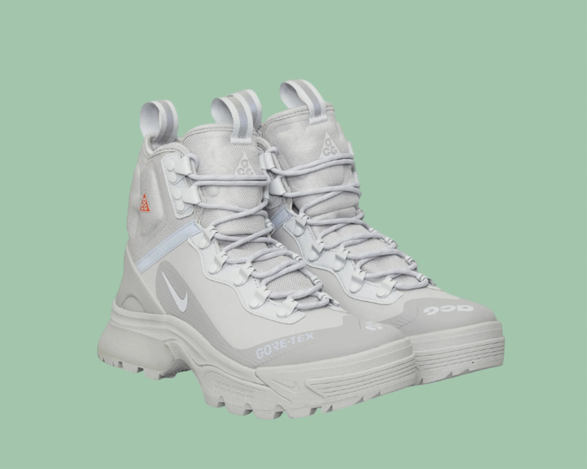 Pair of light gray Nike ACG Gore-Tex hiking boots with thick soles and pull tabs on a green background.