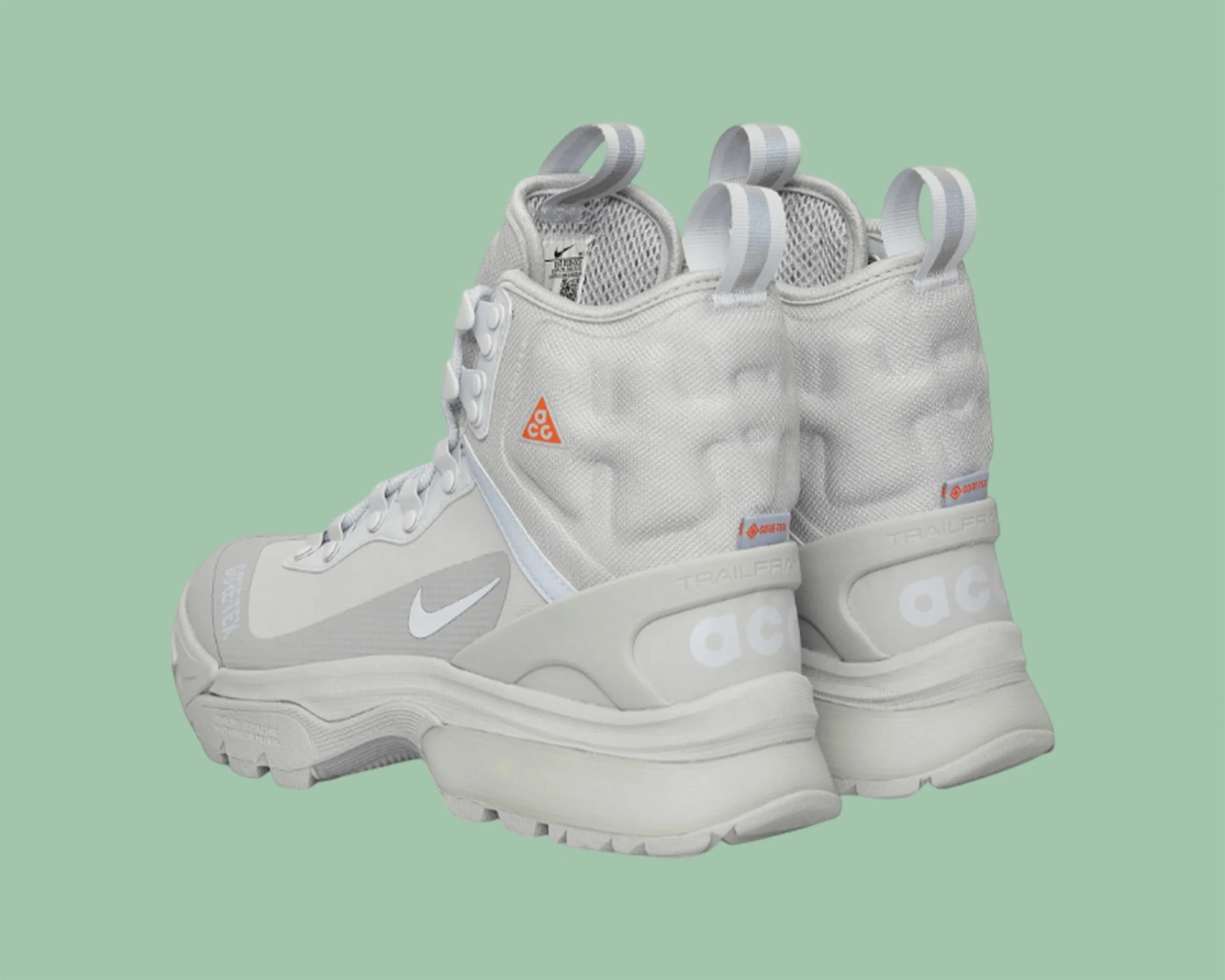 Pair of light gray Nike ACG Trailframe hiking boots with white swoosh logos and orange ACG triangle on a green background.