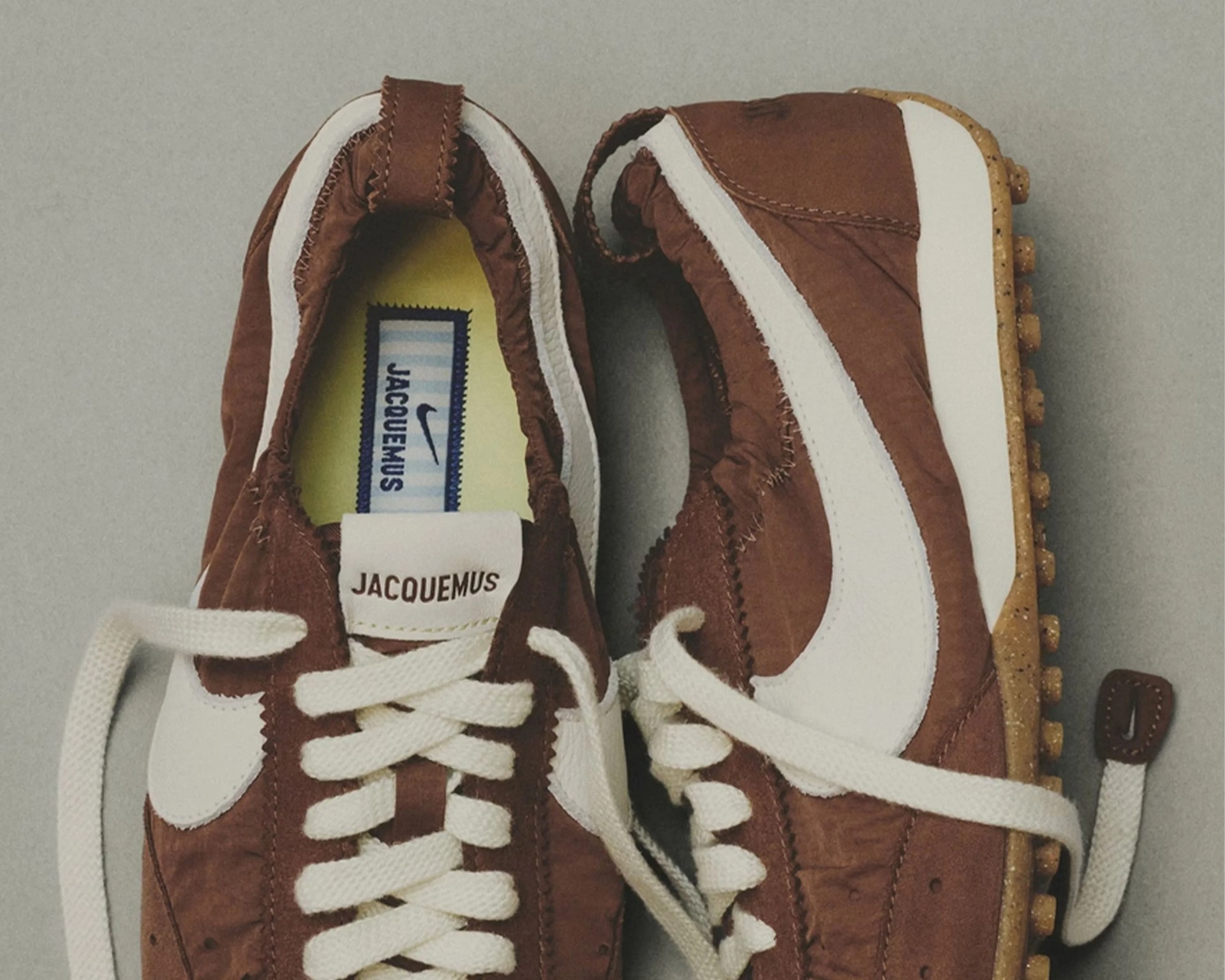 Brown and white Jacquemus x Nike sneakers with white laces and a speckled gum sole on a gray surface.