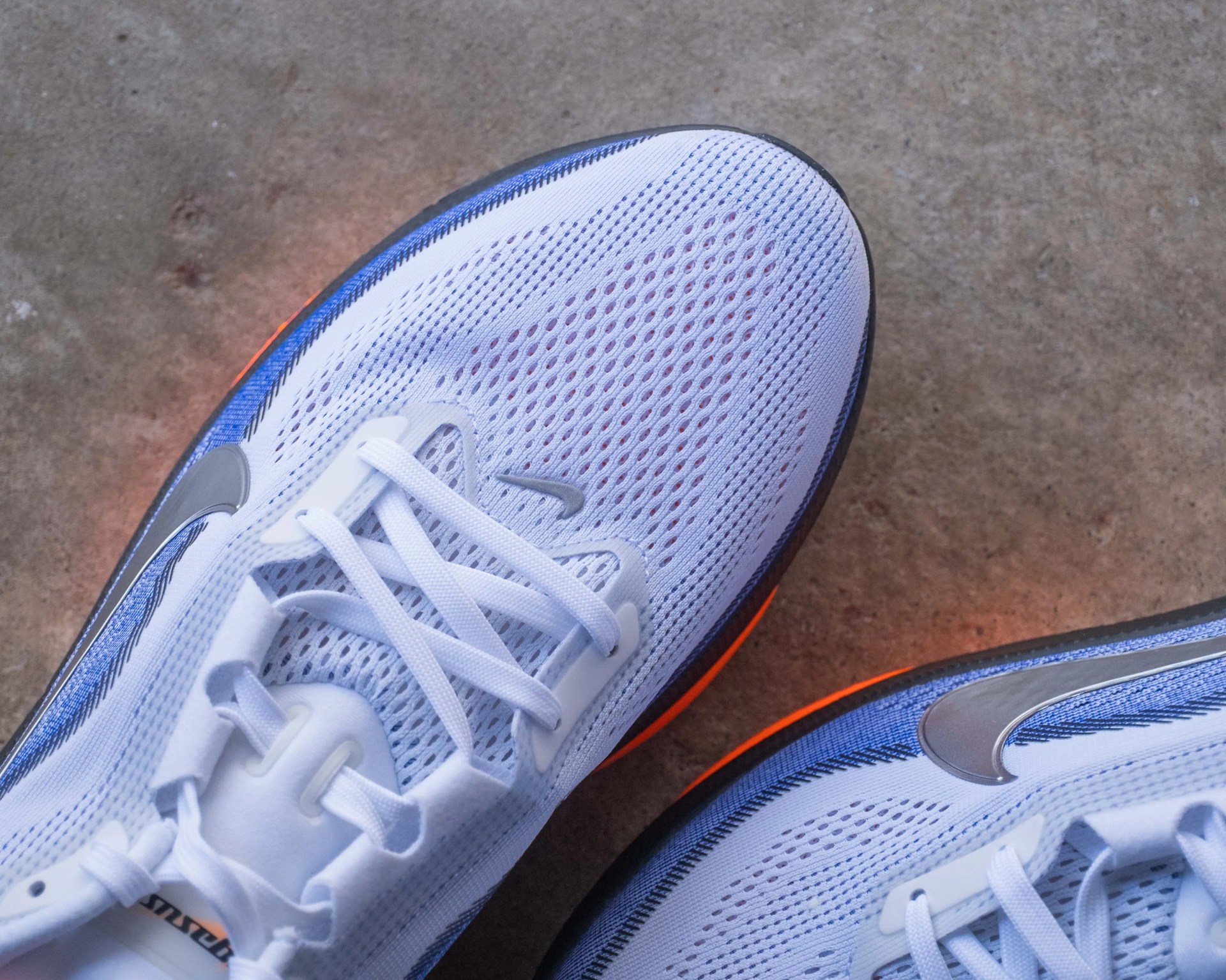 Pair of white Nike running shoes with blue accents and orange soles on a concrete floor.