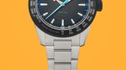 Close-up of a silver metal wristwatch with a black bezel and blue-accented hands on an orange background.