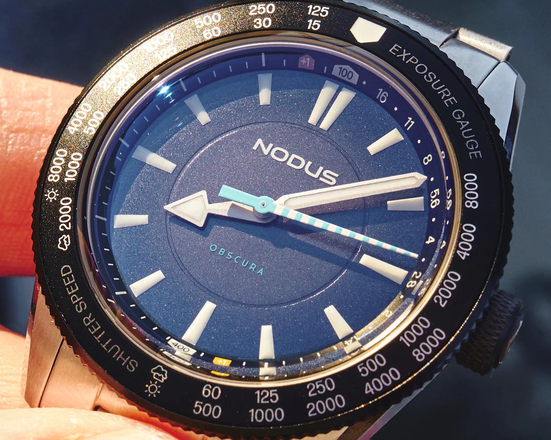 Close-up of a Nodus Obscura watch with a blue dial, white hour markers, and a black bezel marked with shutter speed and exposure gauge scales.