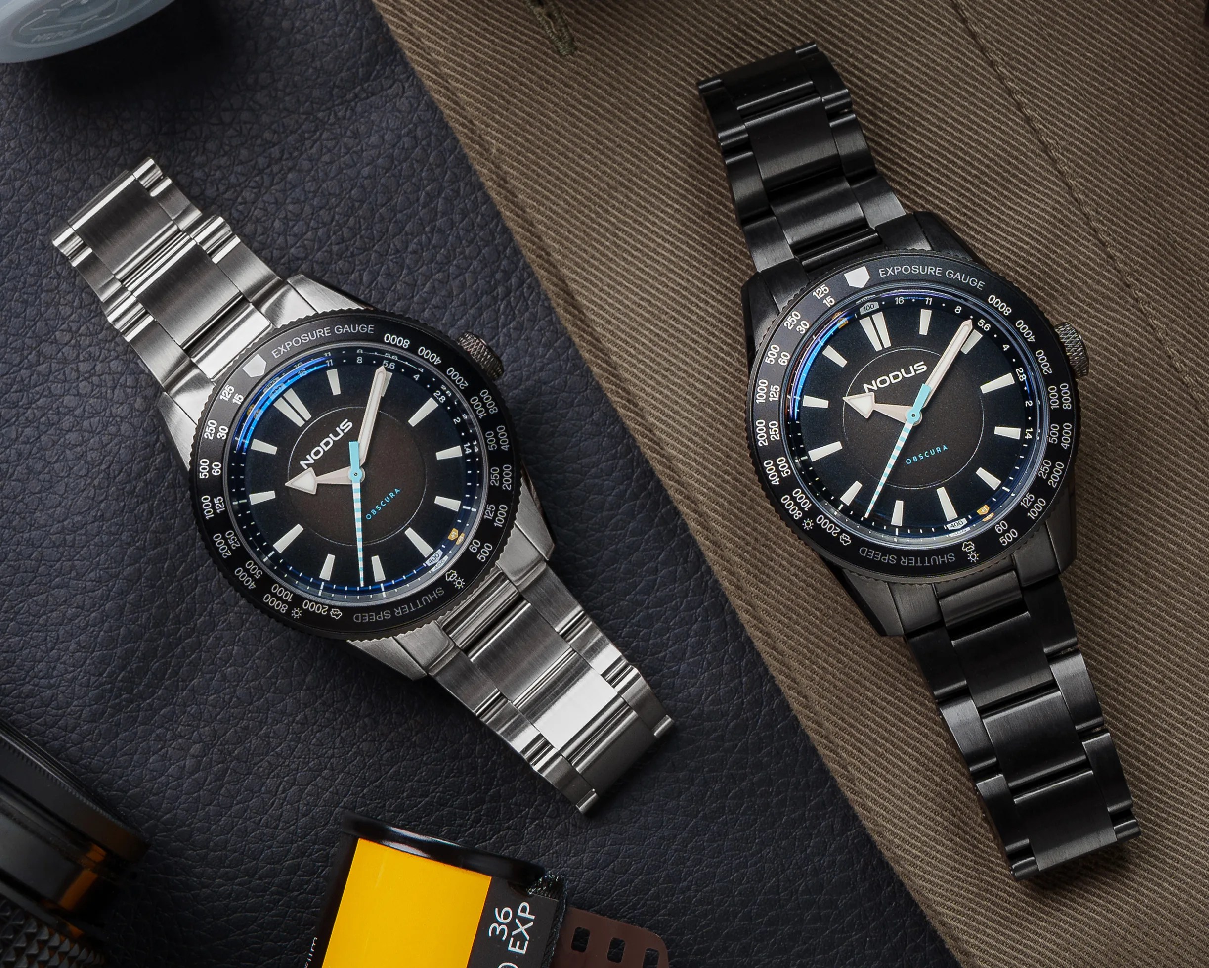 Two Nodus Obscura wristwatches, one with a silver metal bracelet and one with a black metal bracelet, on textured surfaces.