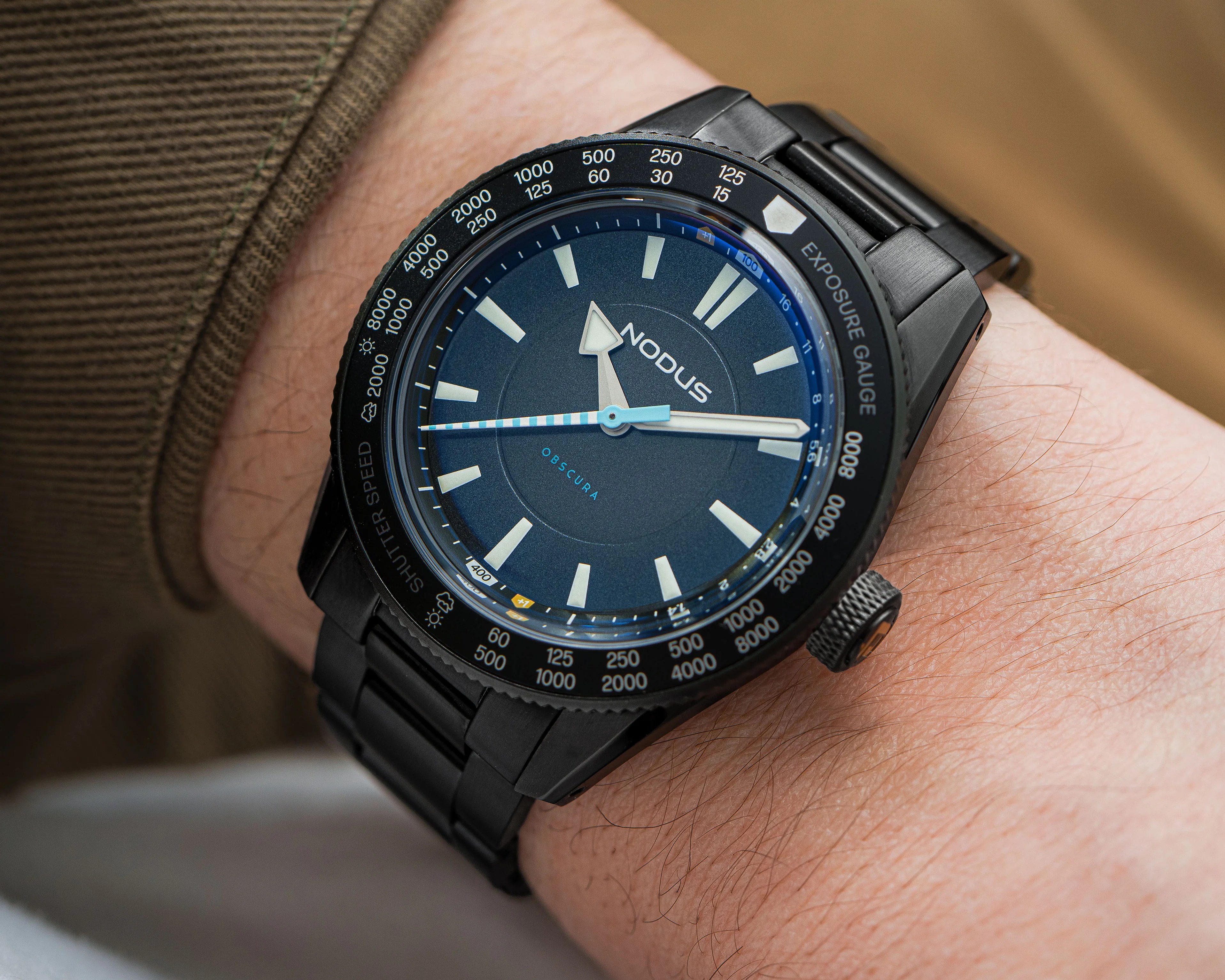 Black metal wristwatch with blue dial and white hour markers, labeled "Nodus Obscura," worn on a wrist.