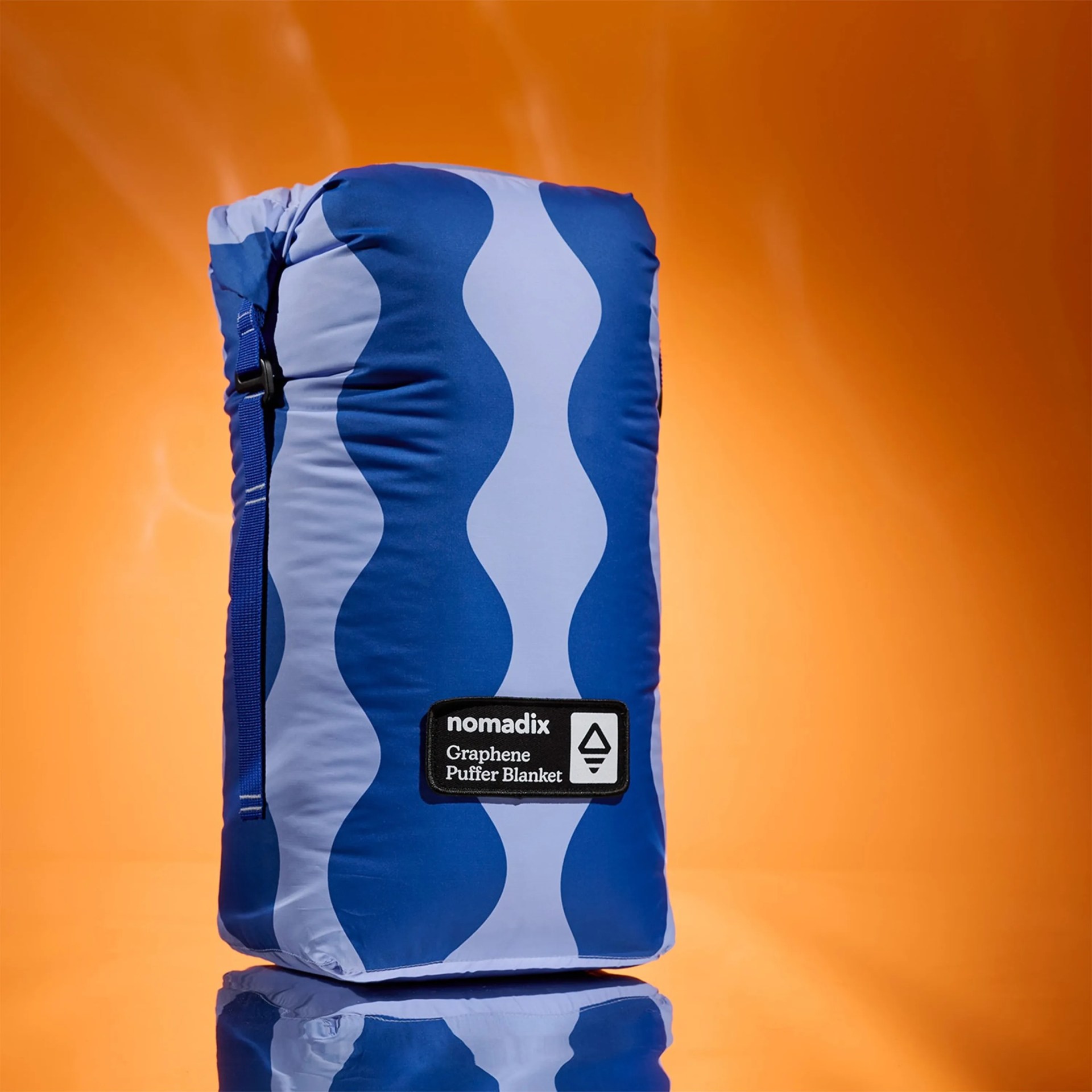 Blue and light blue wavy patterned Nomadix Graphene Puffer Blanket rolled and secured with a strap against an orange background.